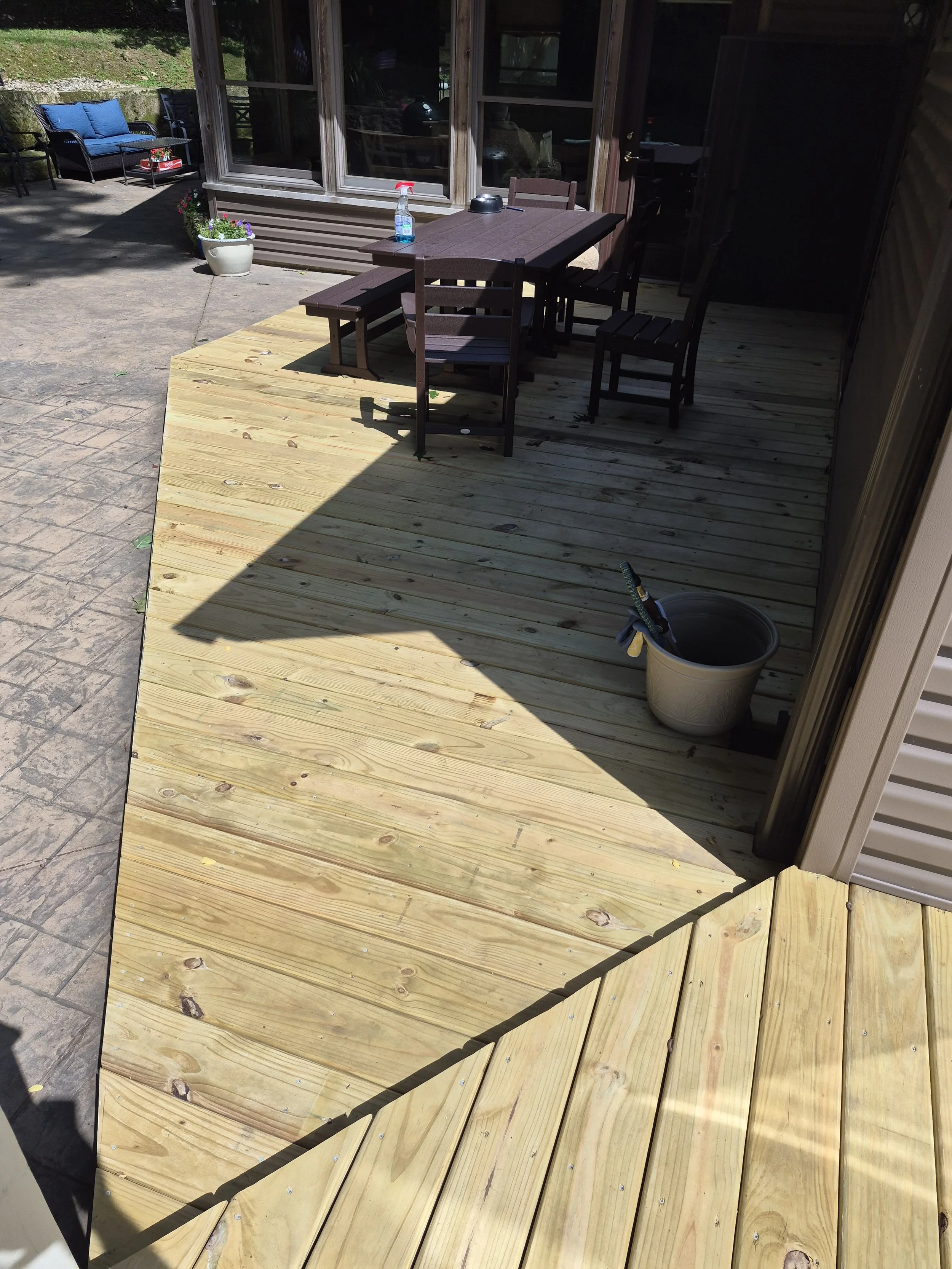 A newly installed wooden deck outside a house with a table, chairs, and a spray bottle on the table. A bucket, some plants, and an outdoor seating area with couches and chairs are visible in the background.