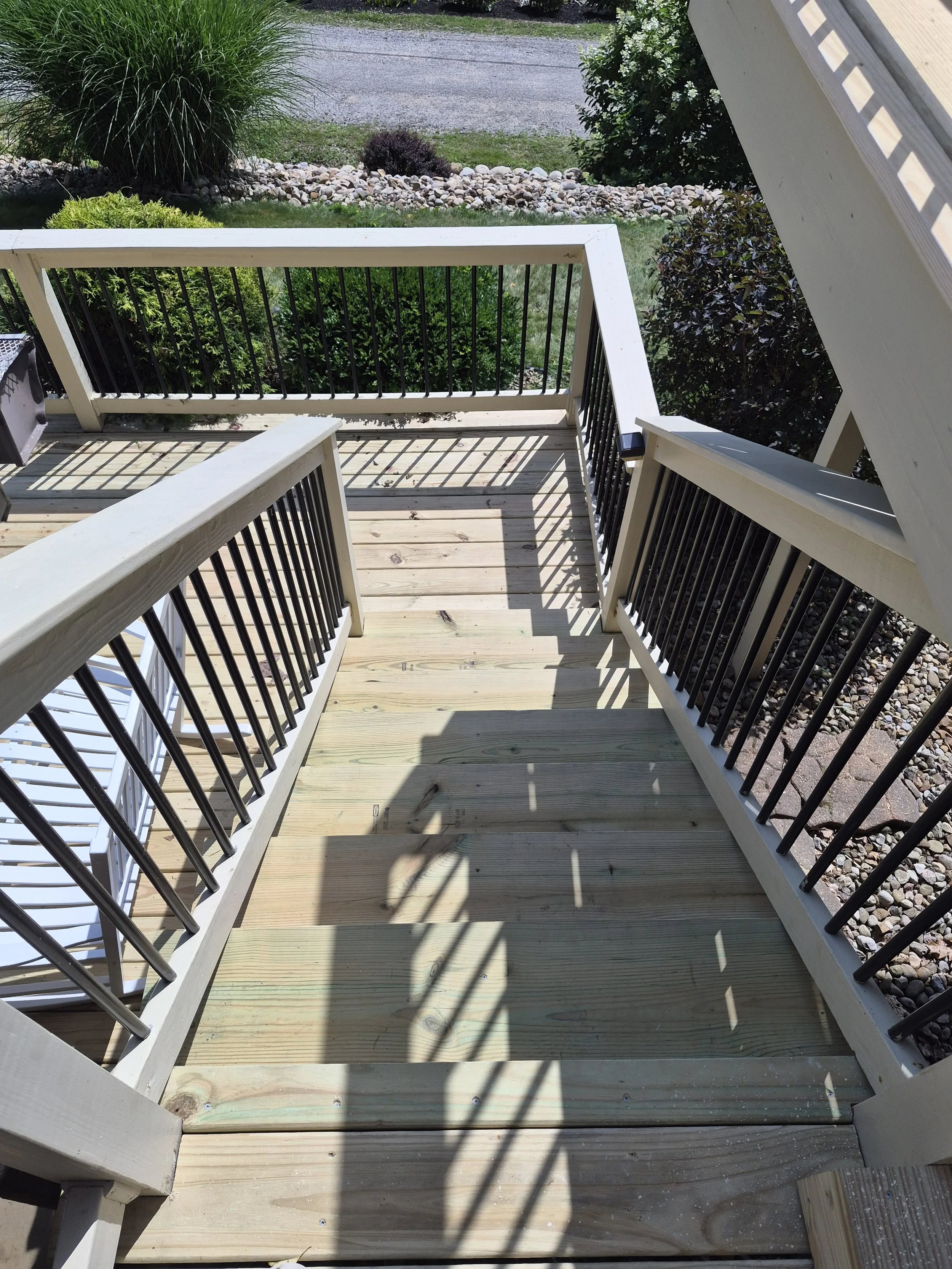 View of a new wooden staircase with black metal balusters leading up to a porch, surrounded by landscaping including bushes, rocks, and a paved road in the background.