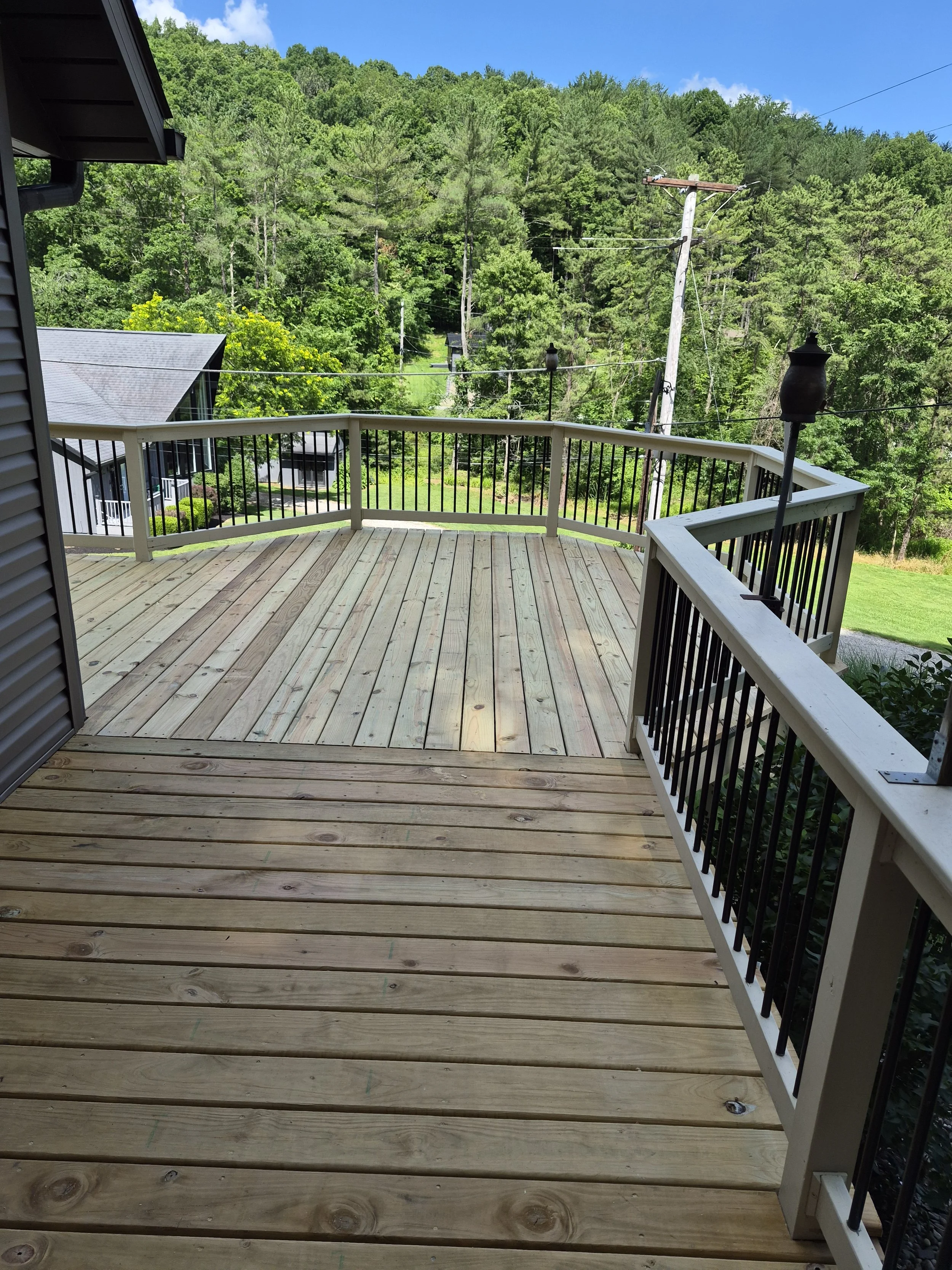 A wooden outdoor deck with railing overlooking a lush, green forested area with trees and hills. A house roof is partially visible on the left side, and utility poles and power lines run through the background under a blue sky with a few clouds.