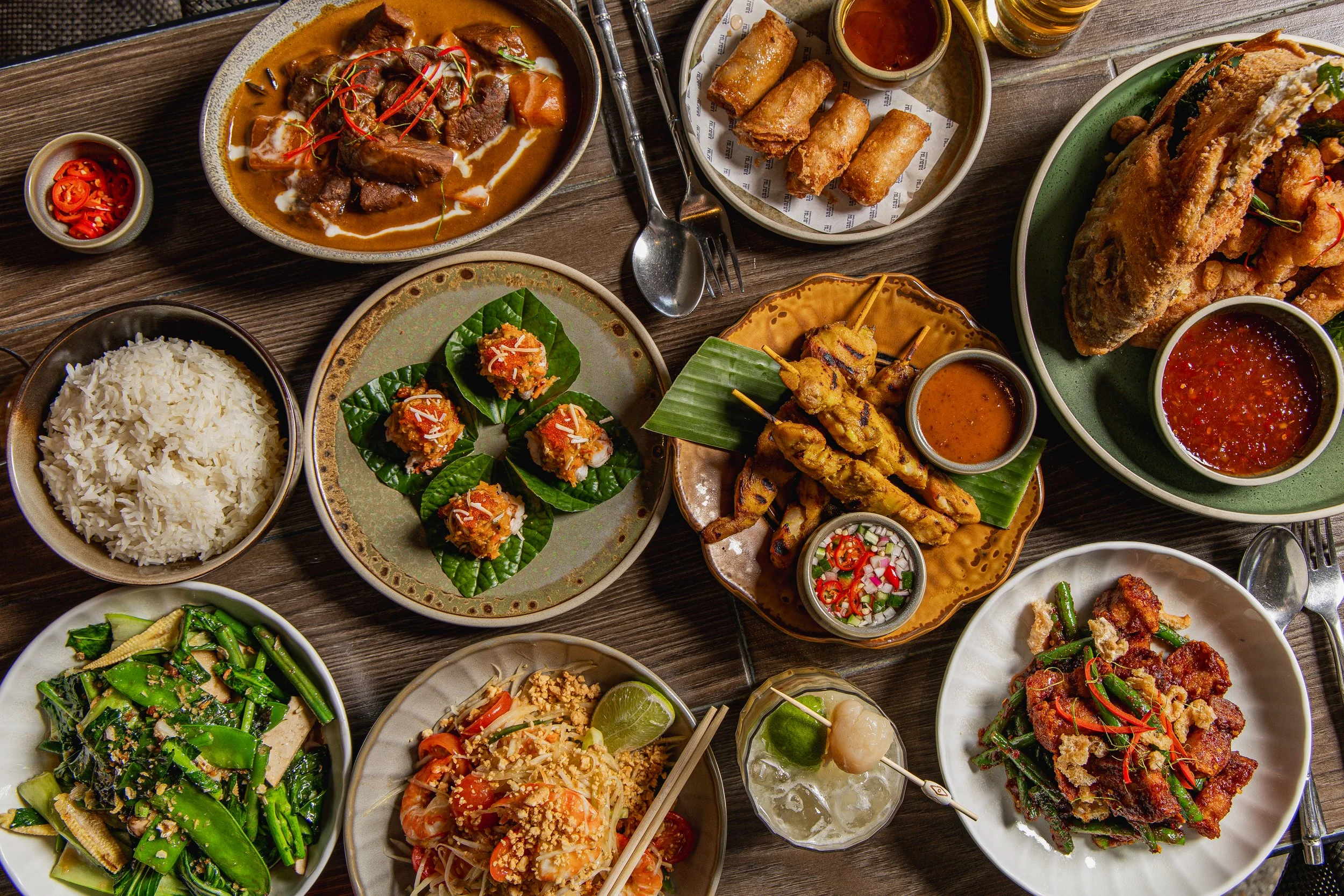 Photo of one of the set menus at Muum Maam, featuring a vibrant spread of Thai dishes including satay skewers, curries, fresh betel leaf bites, spring rolls and stir-fried greens designed for sharing.