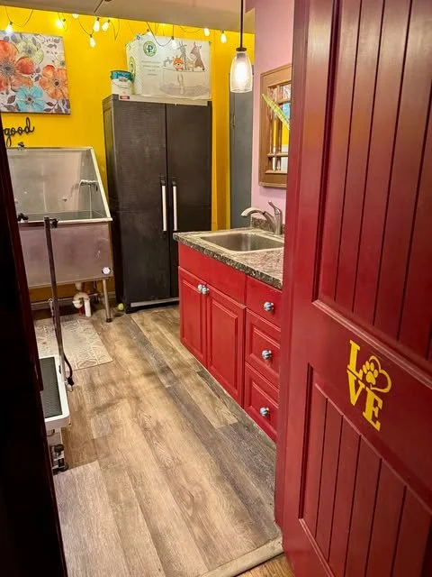 A cozy kitchen with red cabinets, a granite countertop, and a stainless steel sink. There is a black storage cabinet, a stainless steel freezer, and yellow and pink walls decorated with artwork and string lights that spell out "good" and "love."