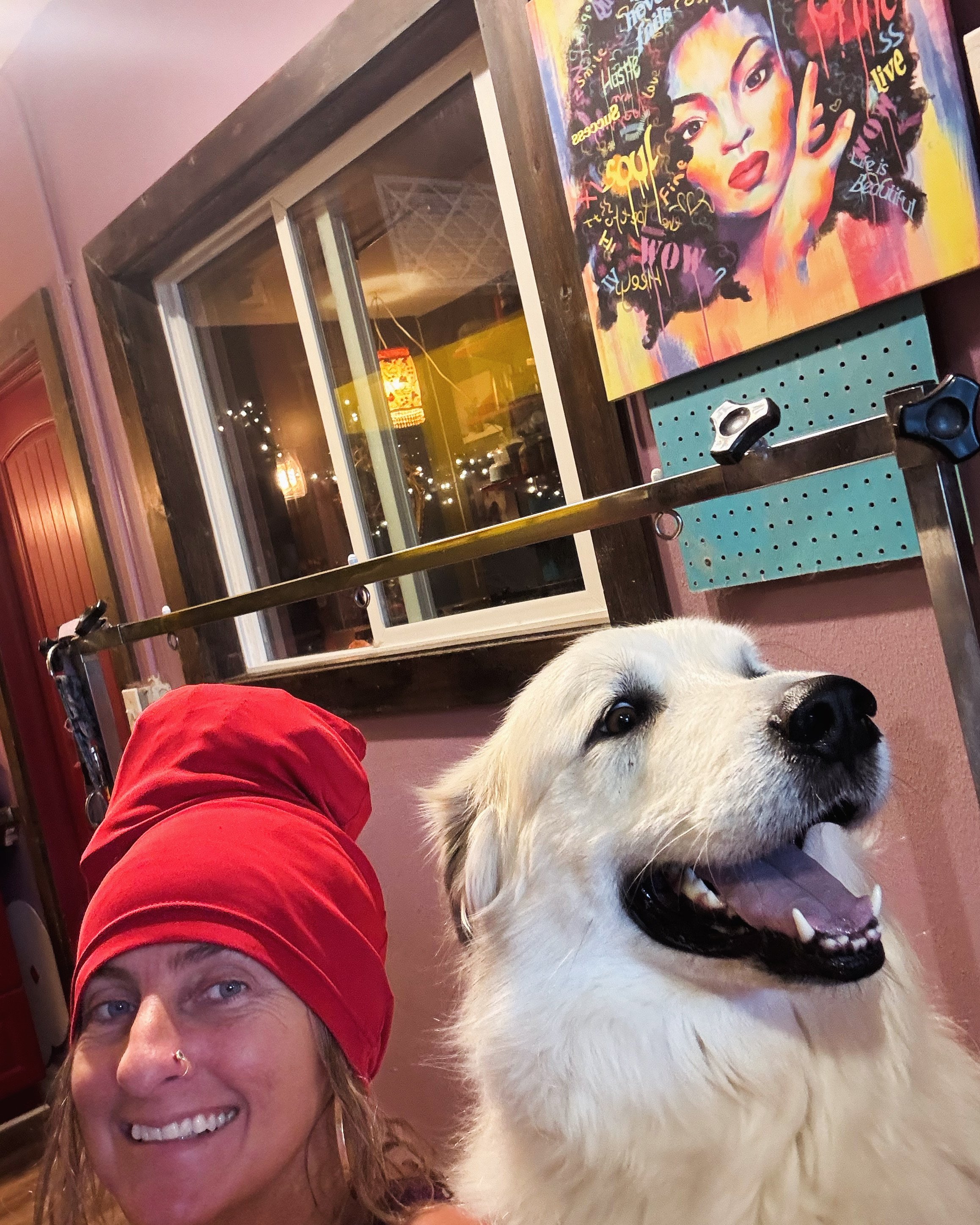 A woman with a red head covering smiling alongside a happy large white dog with a fluffy coat inside a room with colorful artwork and a window in the background.
