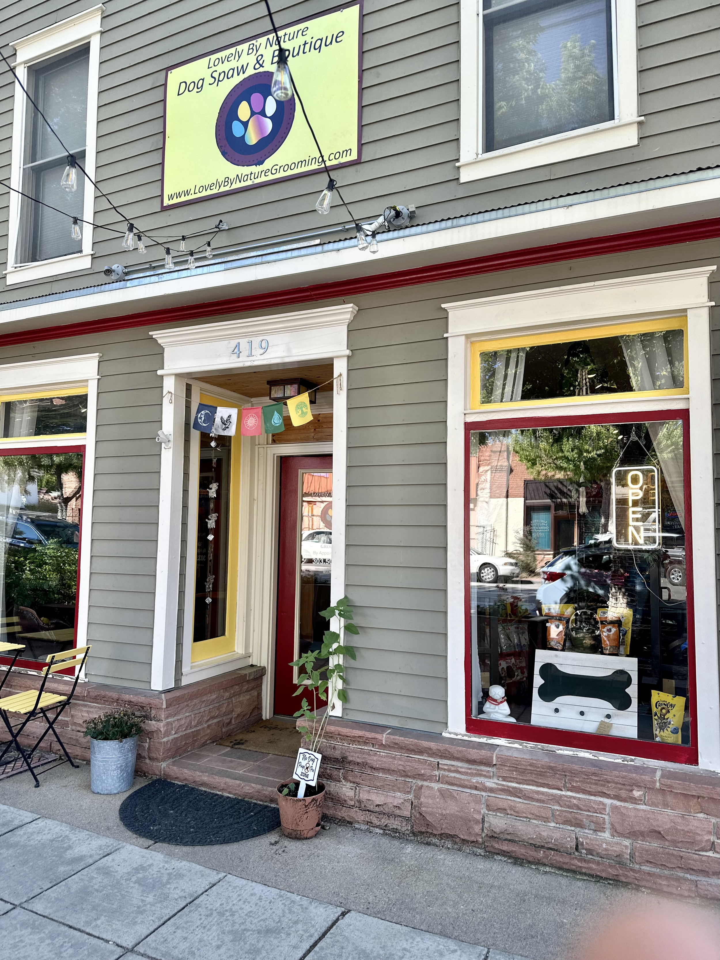 Exterior of a pet grooming business with a neon 'Open' sign in the window, decorative banners, potted plant, and outdoor seating.