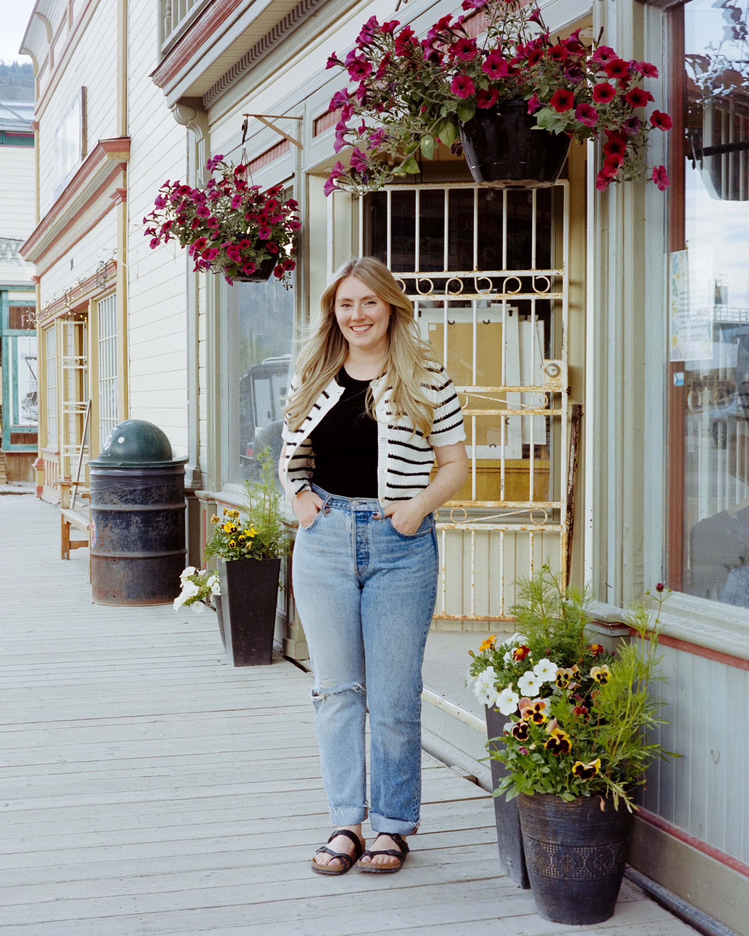 Blonde woman standing on a boardwalk surrounded by flowers in front of her store. She is wearing a white and black stripped cardigan and light blue jeans with rips in the knees.