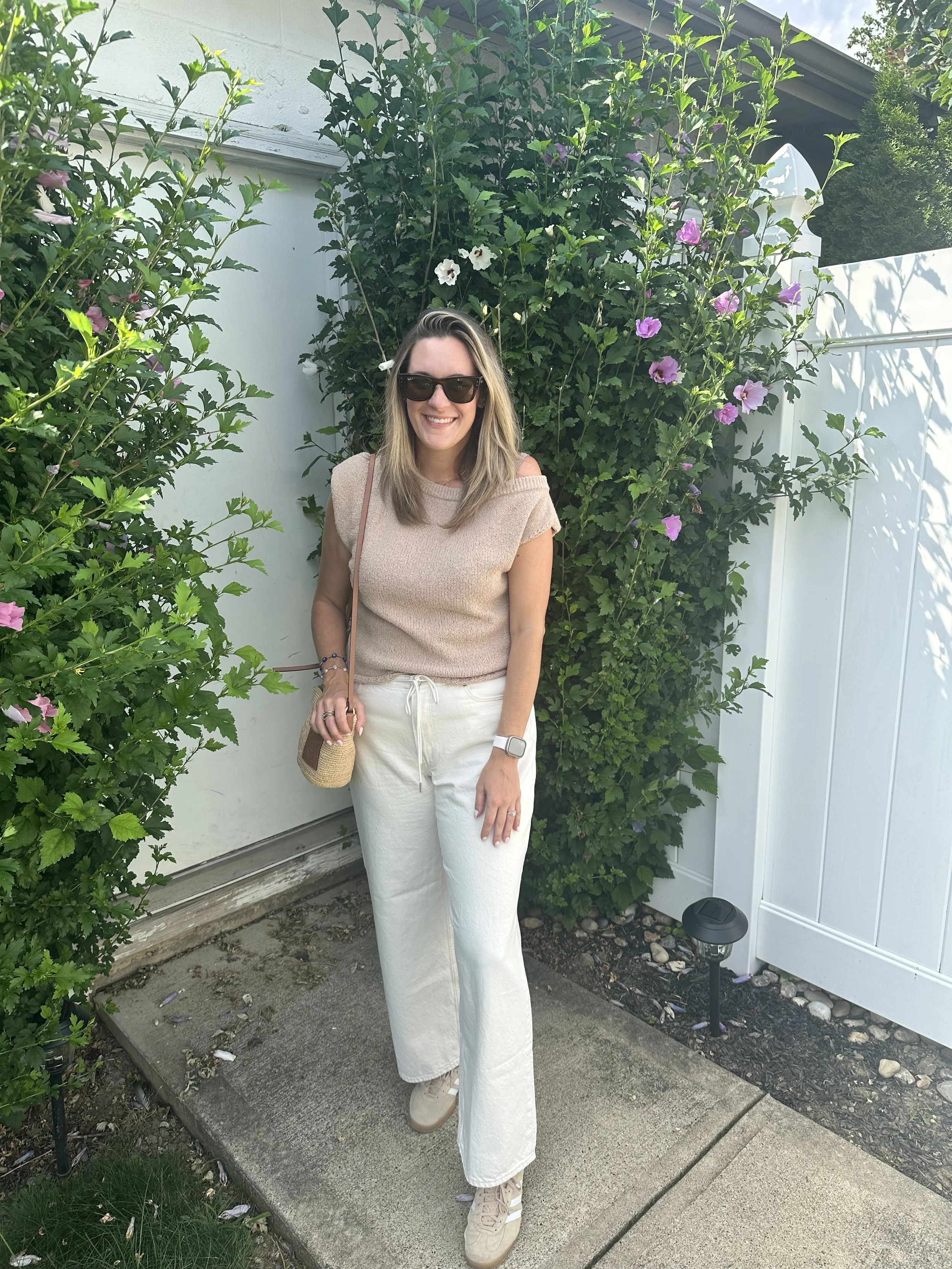 A woman with blonde hair in cream jeans standing in front of flowers and greenery in the summertime.
