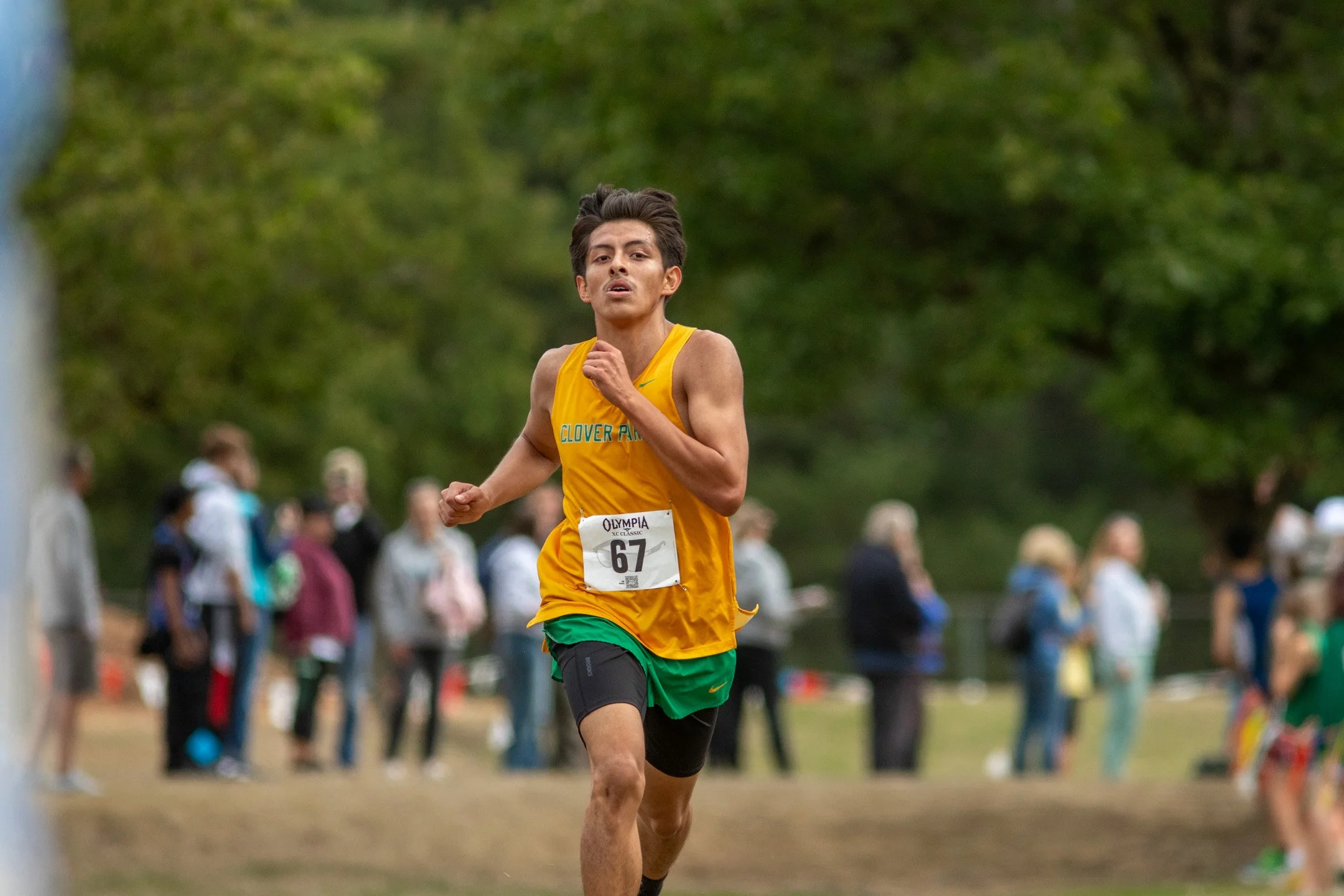Clover Park senior Arturo Baltazar-Magana, bib 67, in a yellow singlet and green shorts, running on a grass course with spectators behind him.
