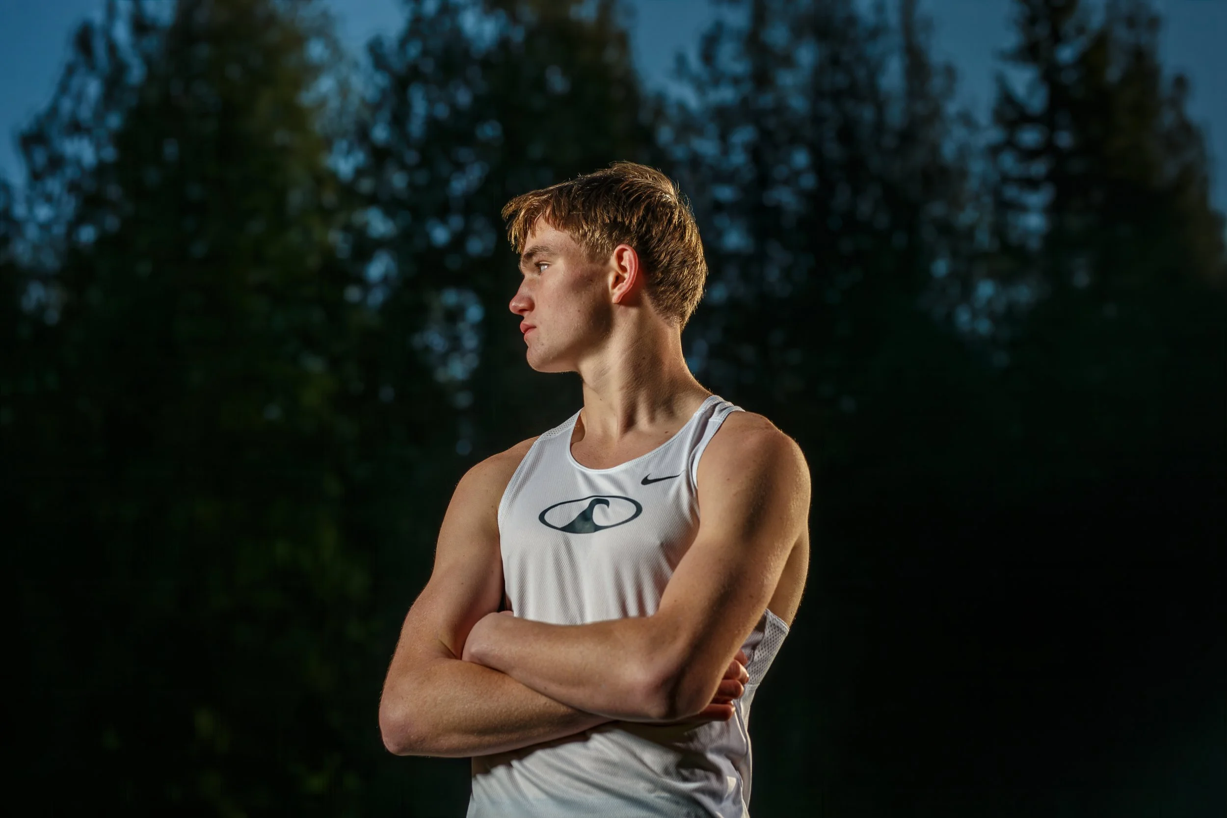 Moody portrait of a male athlete in a white singlet standing against a dark forest background, lit by dramatic rim lighting on his shoulders and hair.
