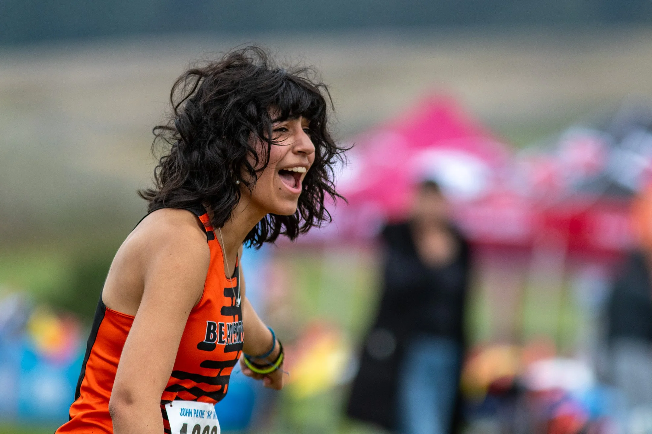 A Beaverton High School runner in an orange and black jersey leaning forward and shouting with an excited smile along the course.