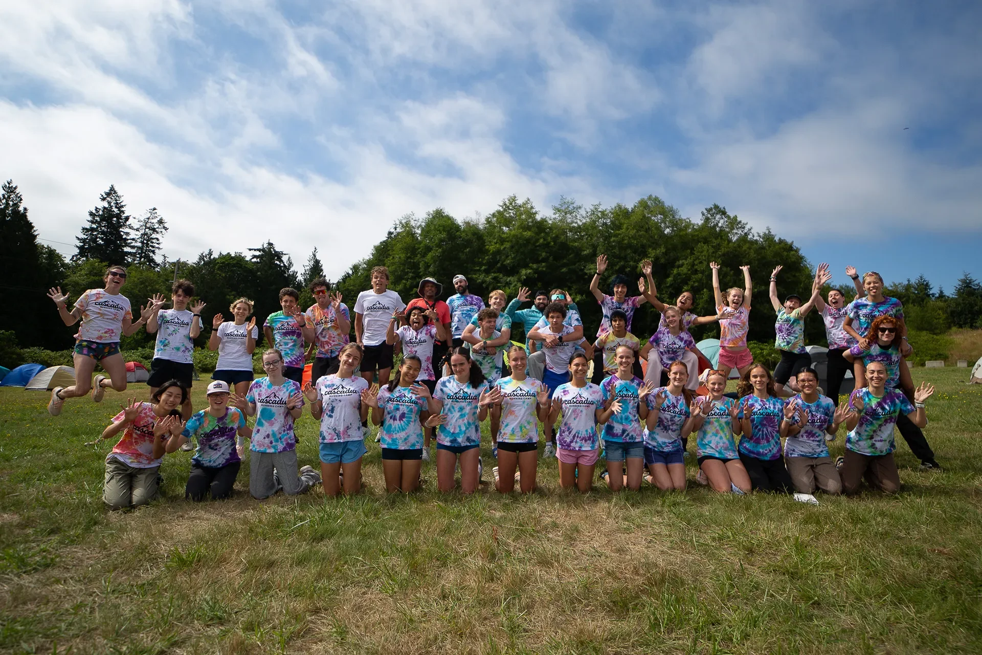 Wide shot of athletes and coaches wearing custom tie-dye shirts cheering and throwing their hands up for a final group photo at camp.