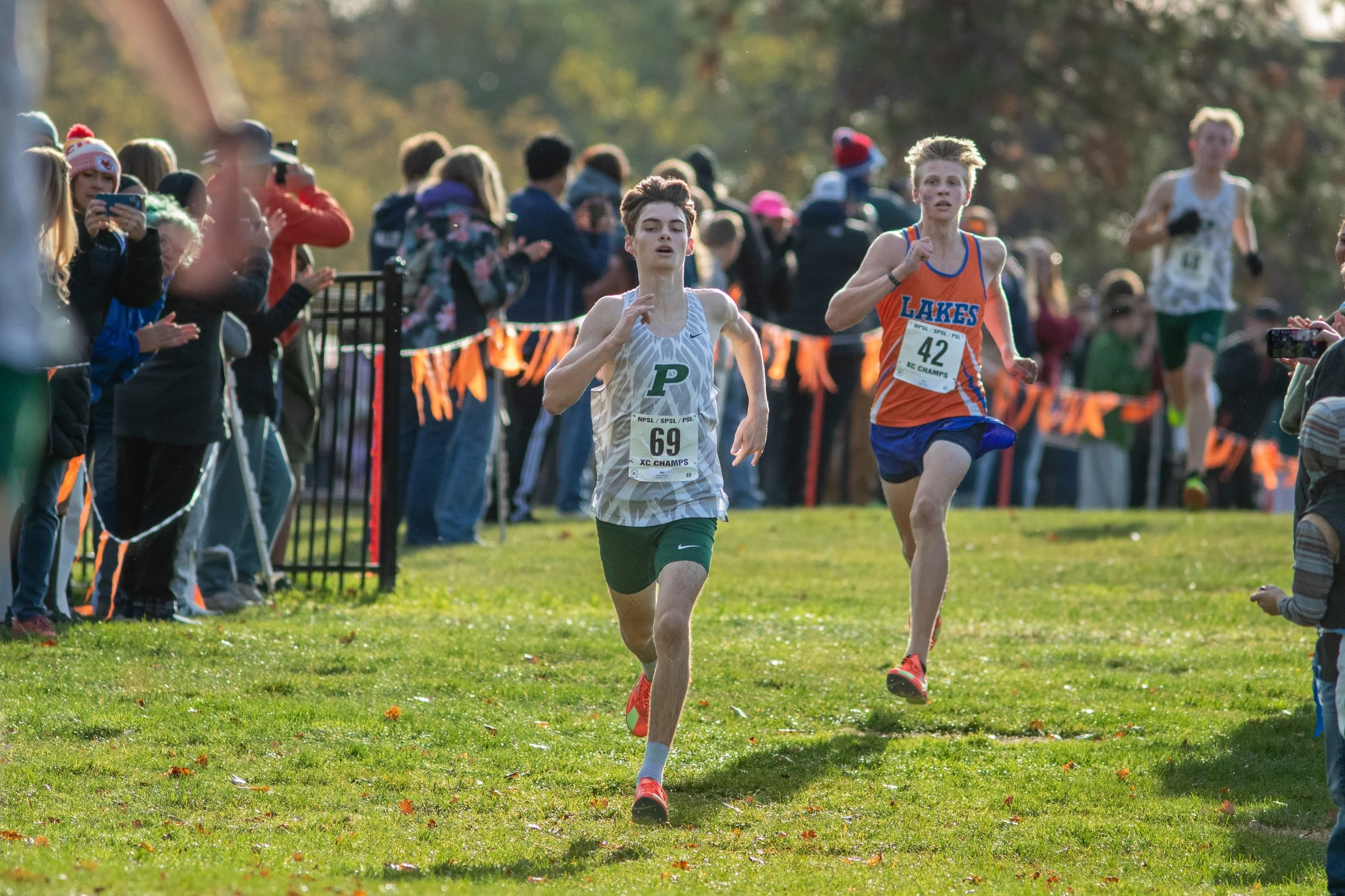 Nolan Winderl of Peninsula, bib 69, in a green and white singlet, runs on grass ahead of a Lakes runner in bib 42 as spectators line the orange-flagged course.