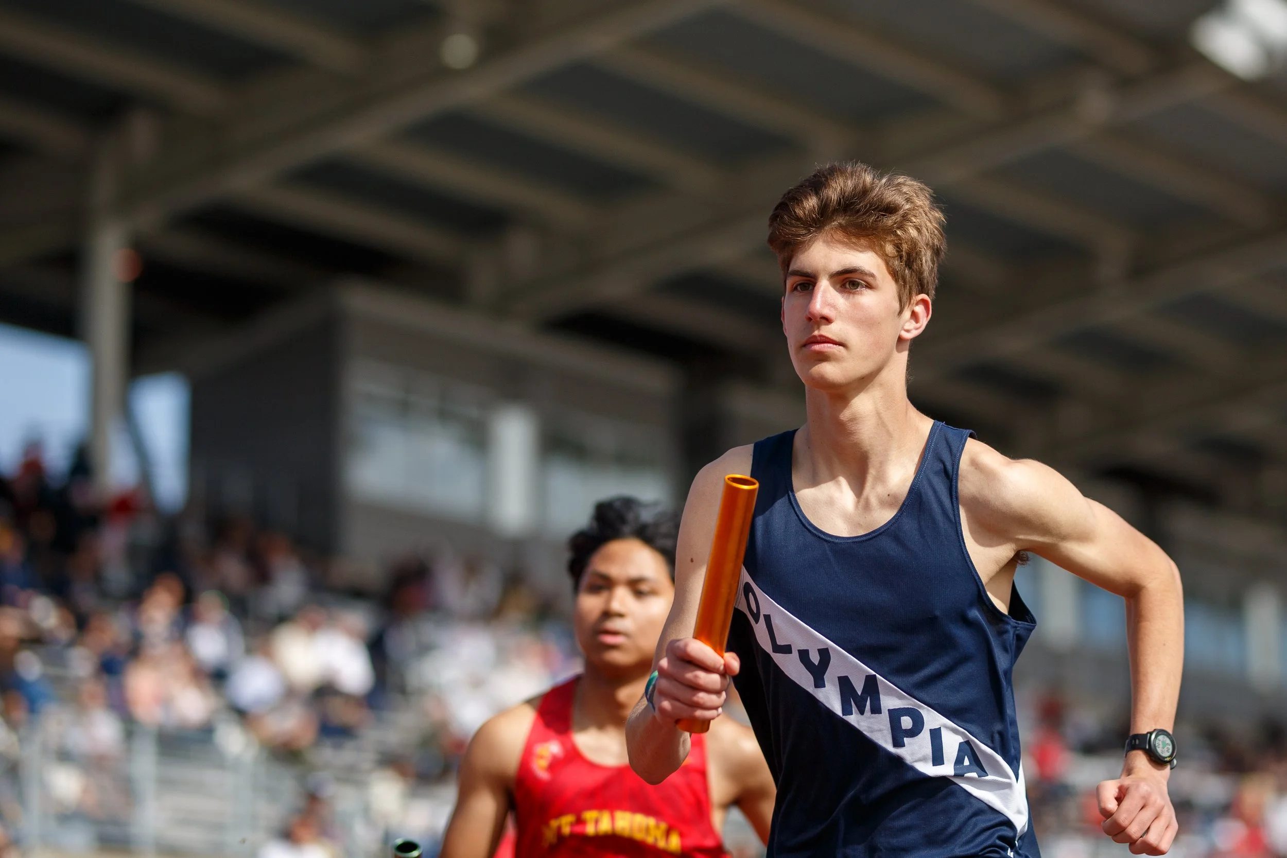 Theodore Ruder of Olympia High School running with a relay baton, wearing a navy Olympia singlet, with a competitor in a red uniform behind him.