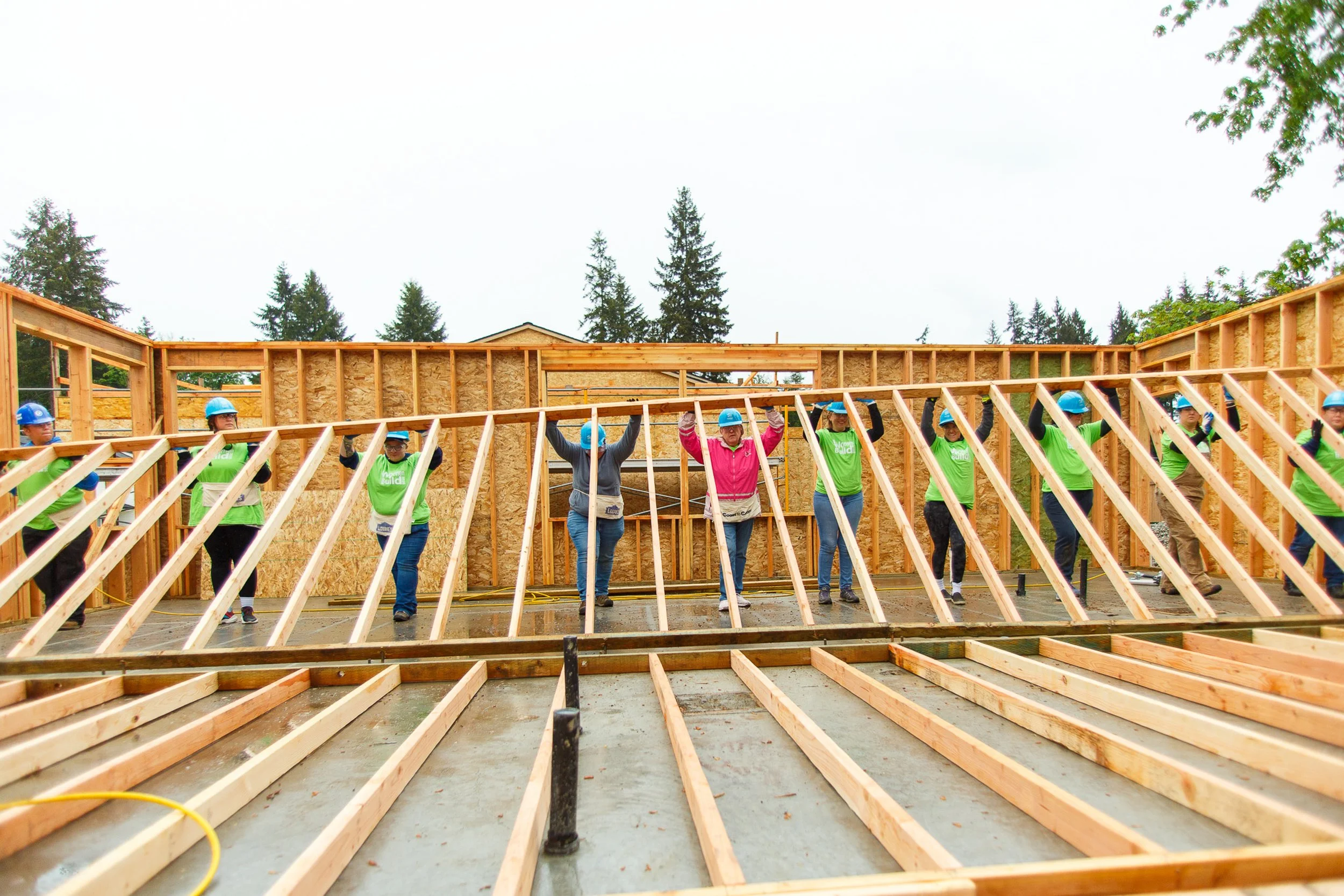 A large group of volunteers working together to manually lift a large wooden wall frame into a vertical position on a concrete foundation.