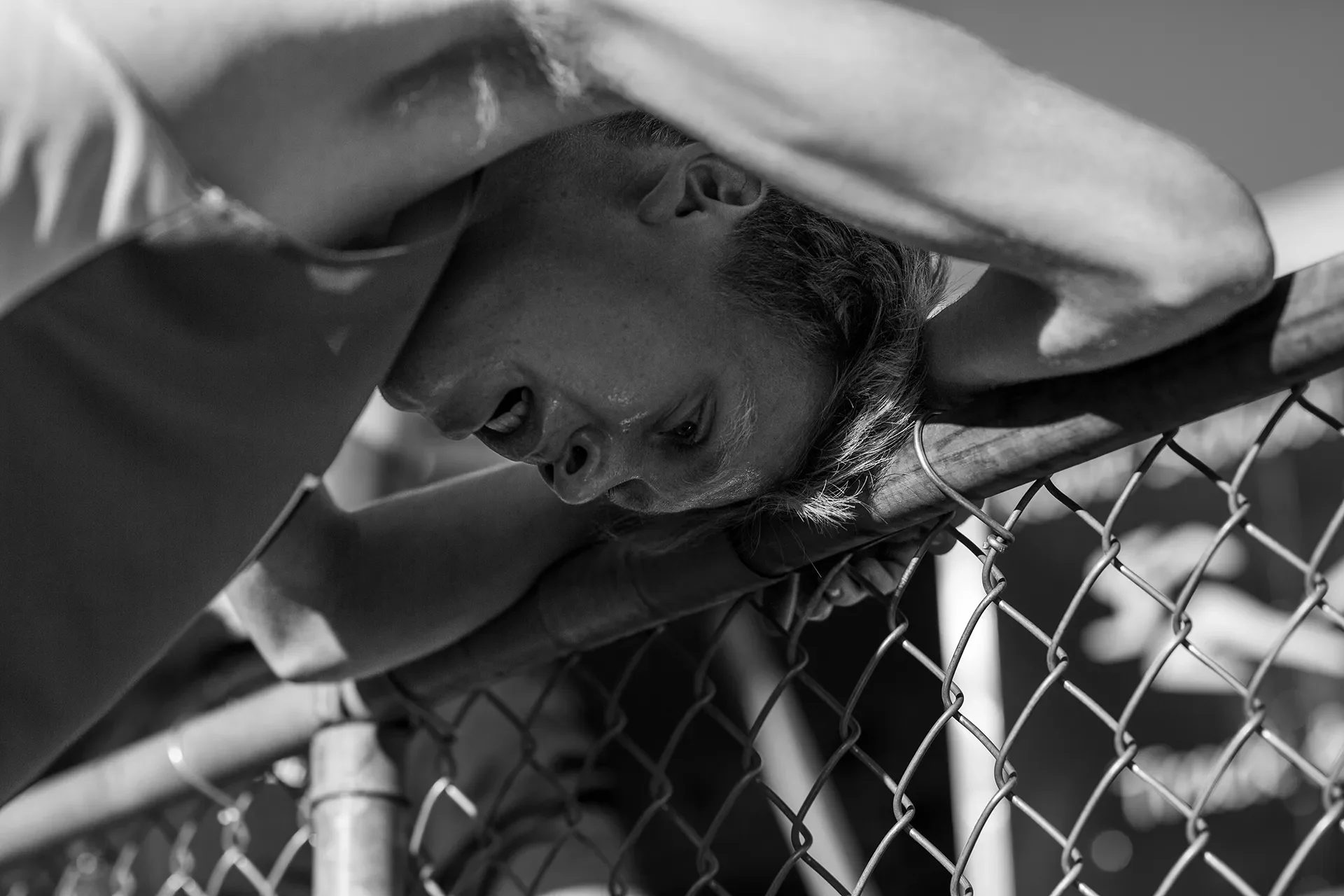 A gritty black and white close-up photograph of Camas high school distance runner Cohen Butler resting his head and arms heavily on a top rail of a chain-link fence, looking completely physically exhausted after a race.