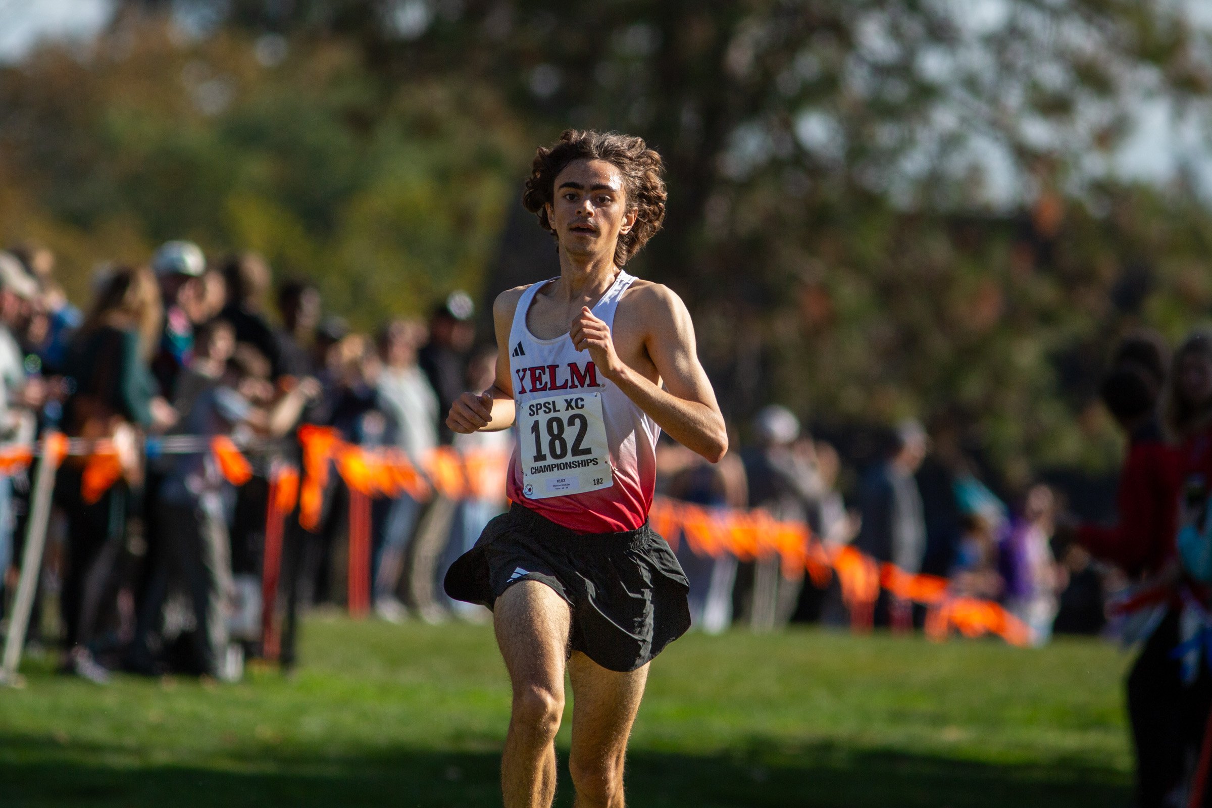 Marcos Andujar of Yelm, bib 182, runs on grass in a white and red Yelm singlet and black shorts, with spectators behind orange course flags.