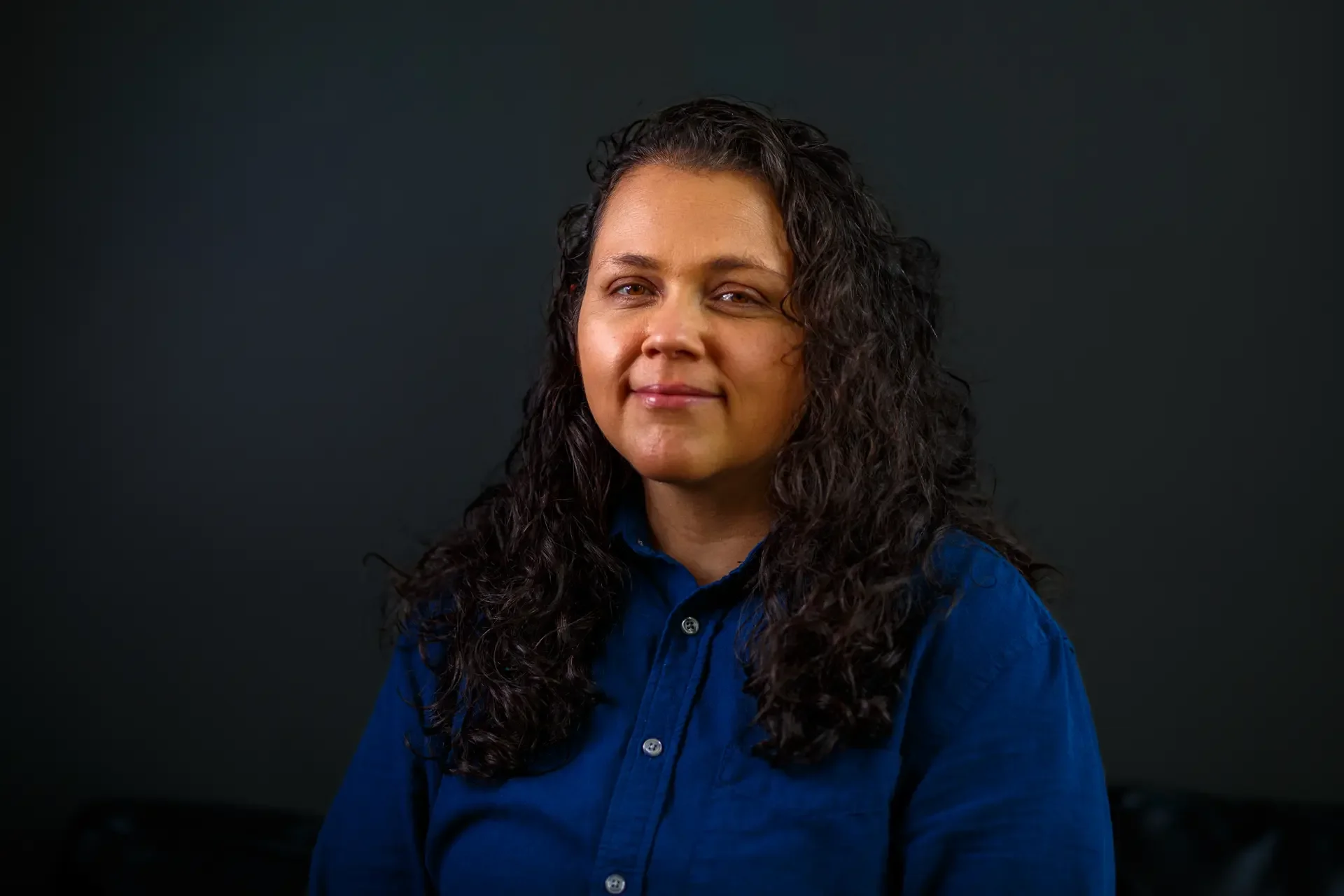 Consistent corporate headshot of a woman in a blue shirt against a dark background, showing unified team photography styling.