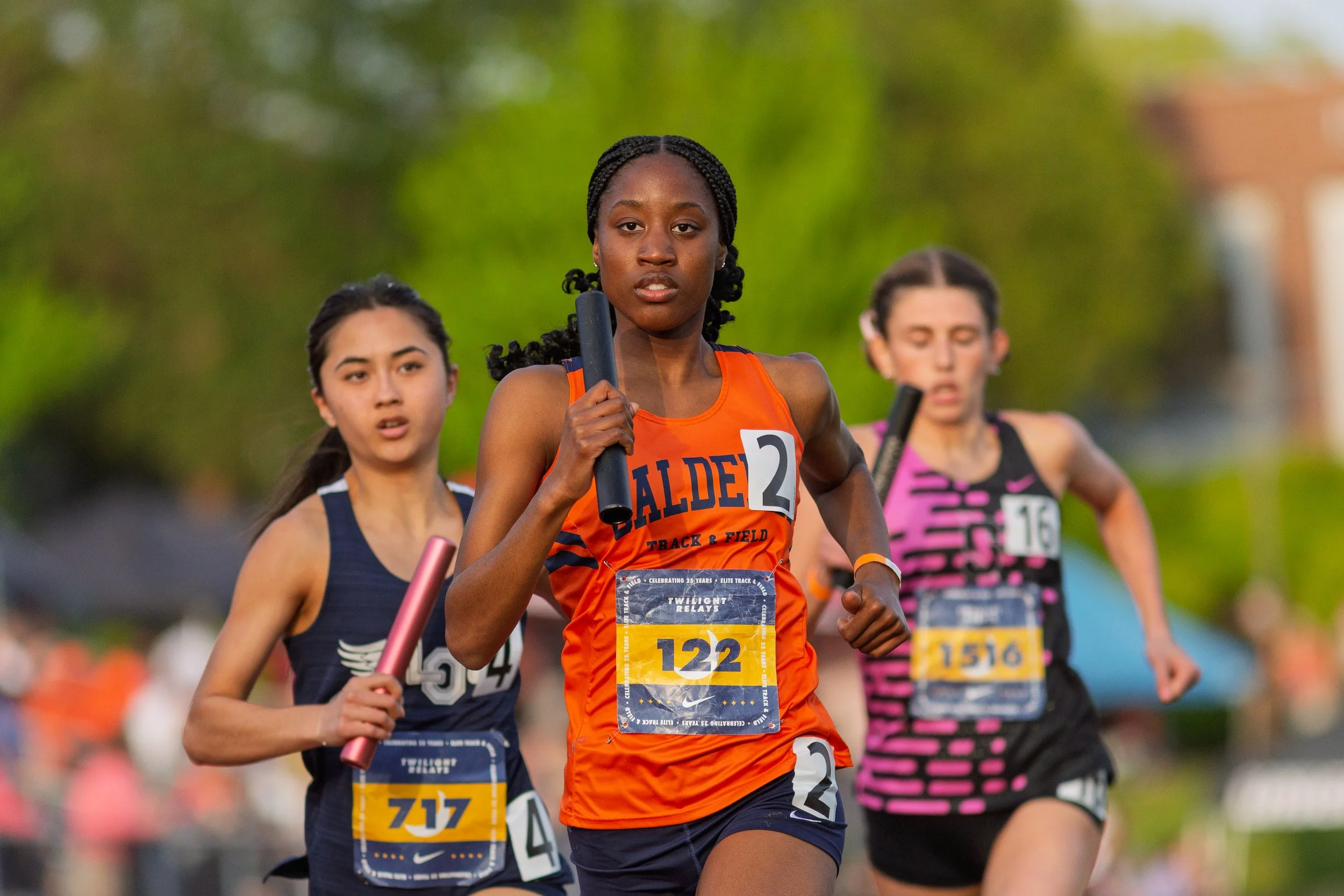 Athlete in orange jersey sprints during Twilight Relays track and field event while holding black baton. Competitive runners follow closely behind during intense relay race competition.