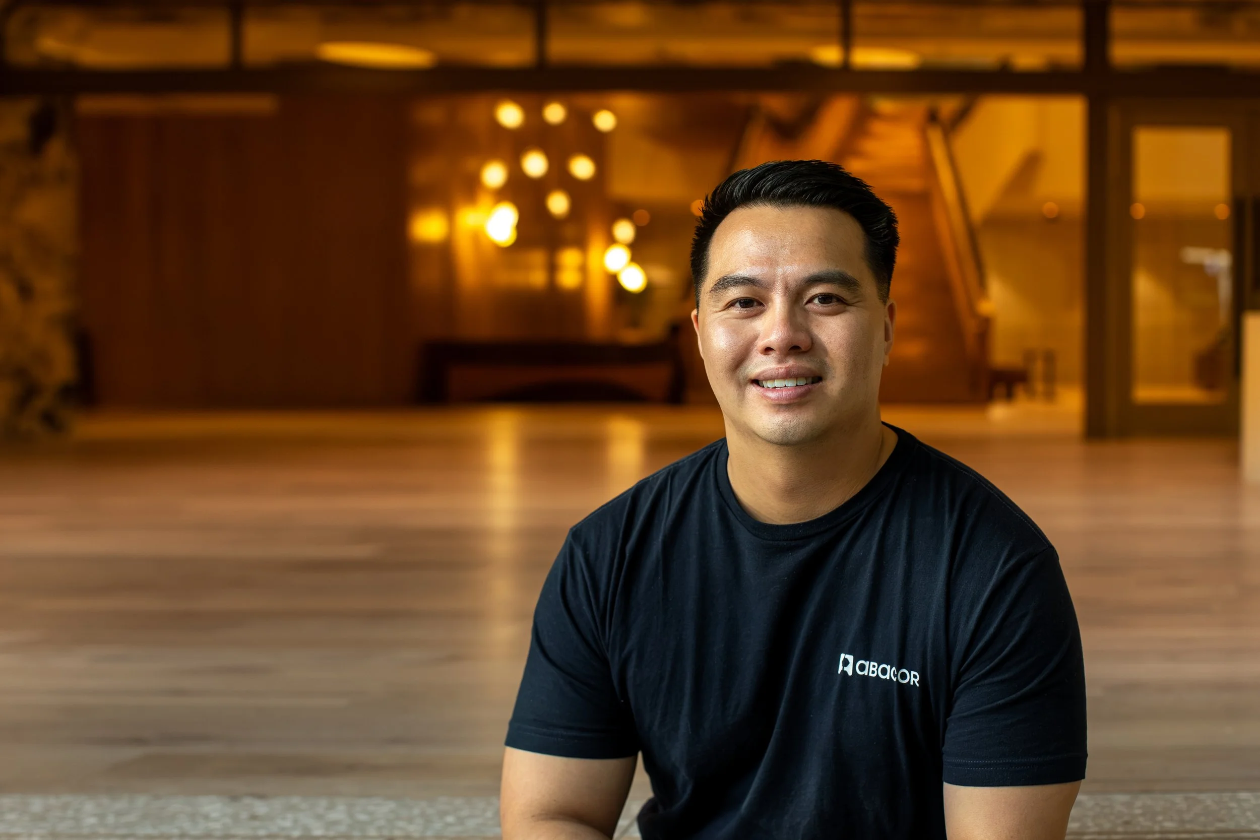 A man with short black hair and a black T-shirt with the logo 'ABACOR' on it, smiling and sitting indoors with a wooden staircase and warm yellow lighting in the background.