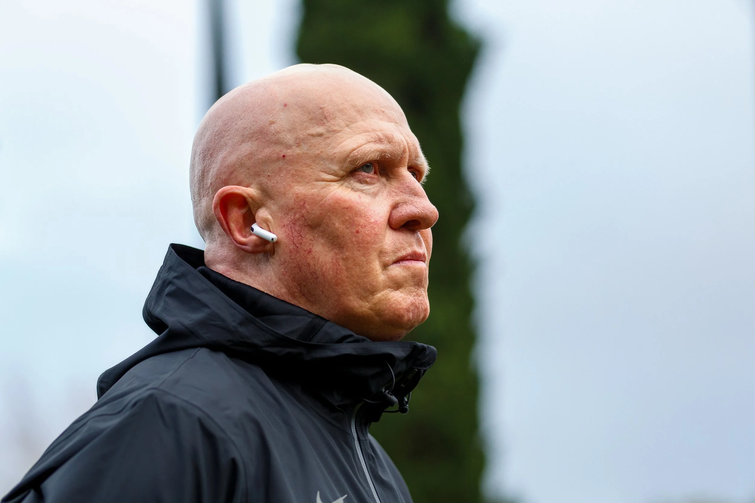 A close-up profile portrait of Tom Rothenberger, the Jesuit High School track and field coach, wearing a black Nike jacket and white earbud during an event in Portland, photographed by Russell Moore.