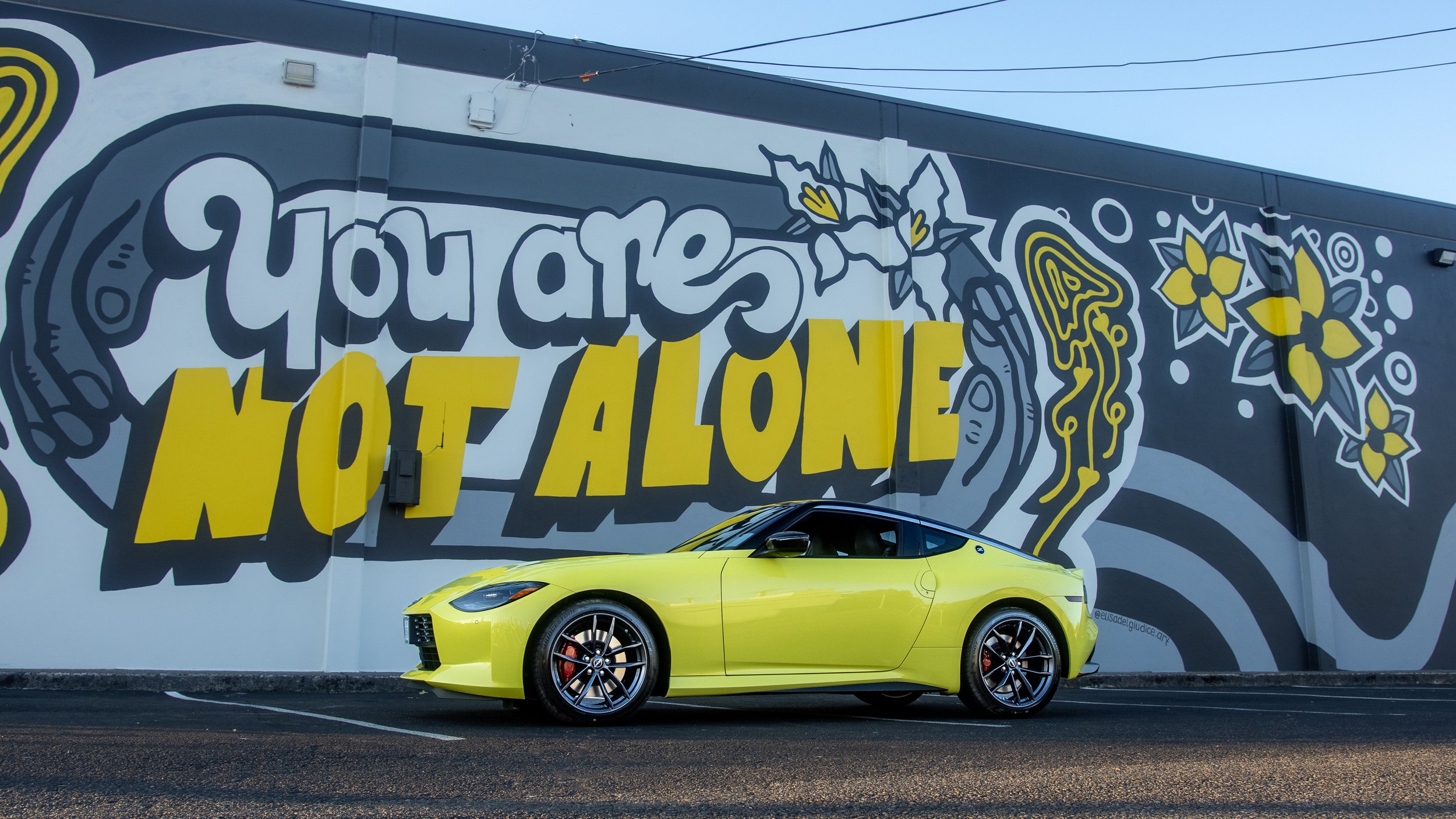 A bright yellow Nissan Z sports car parked in front of the finished "You Are Not Alone" mural in Olympia, WA, matching the yellow lettering perfectly.