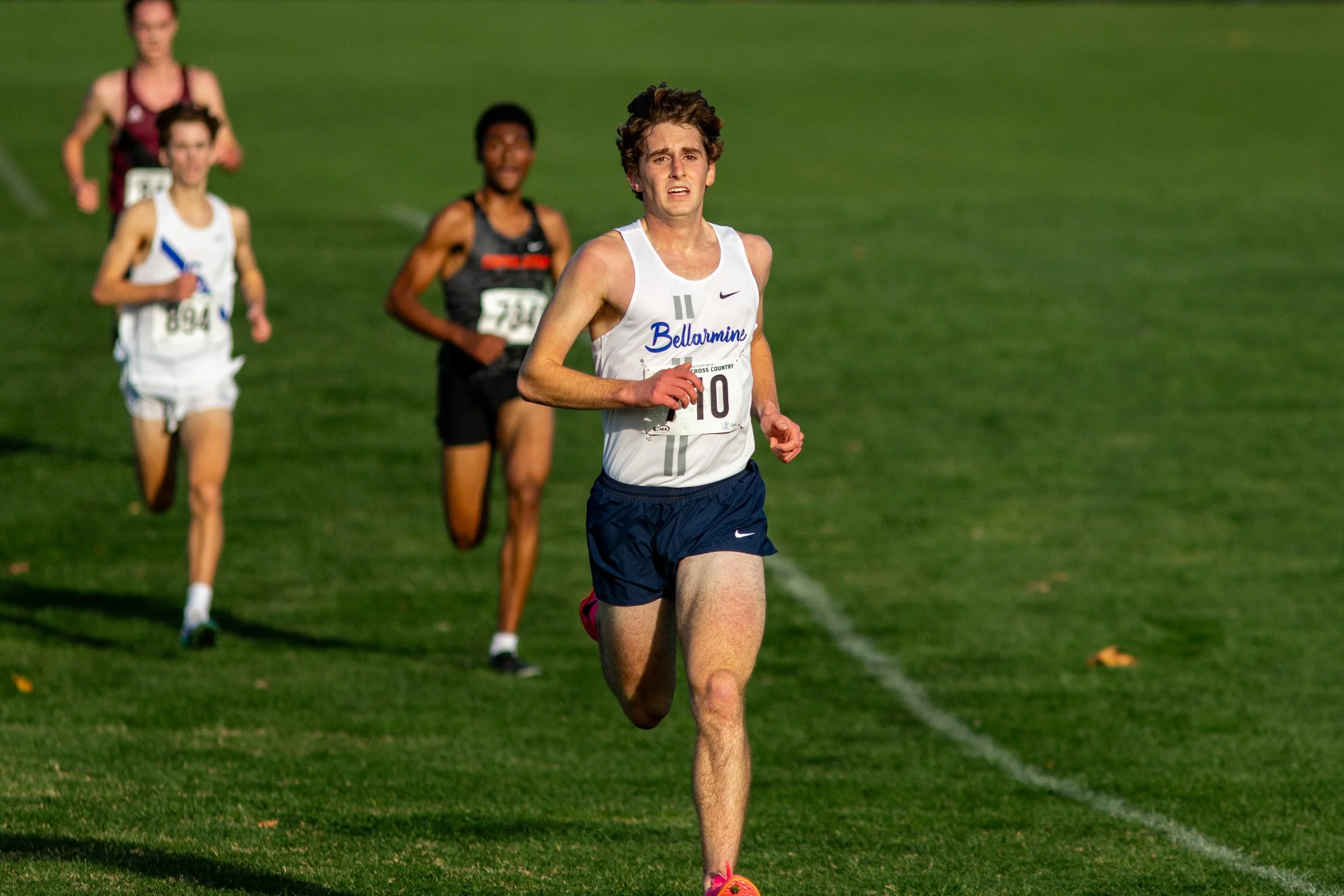 Luke Saalfeld of Bellarmine Prep in a white Bellarmine Prep singlet with navy script lettering, bib 710, leading a group of runners across a green grass field with a crowd along the far edge.
