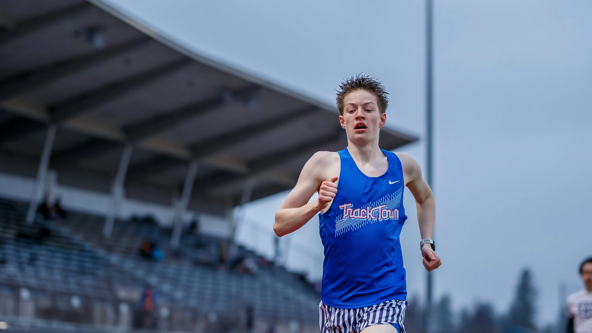 Male athlete wearing blue Track Town singlet sprints on outdoor track. Competitive runner shows intense focus during race near stadium bleachers under cloudy sky. Professional athletic event.