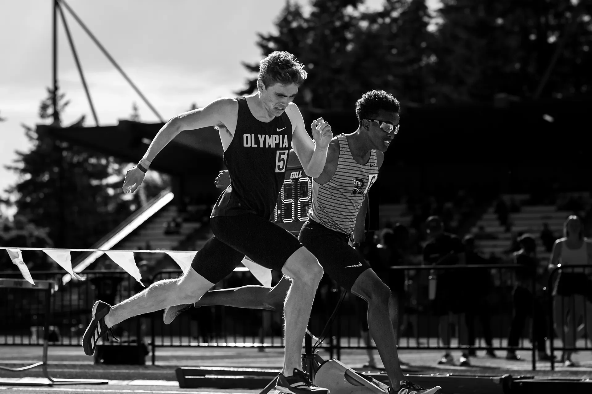 A high-contrast black and white photograph of two male track athletes sprinting in a dead heat. On the left, Olympia's Caden Durocher in a dark singlet lunges forward shoulder-to-shoulder with Rogers' Amin Fidaar