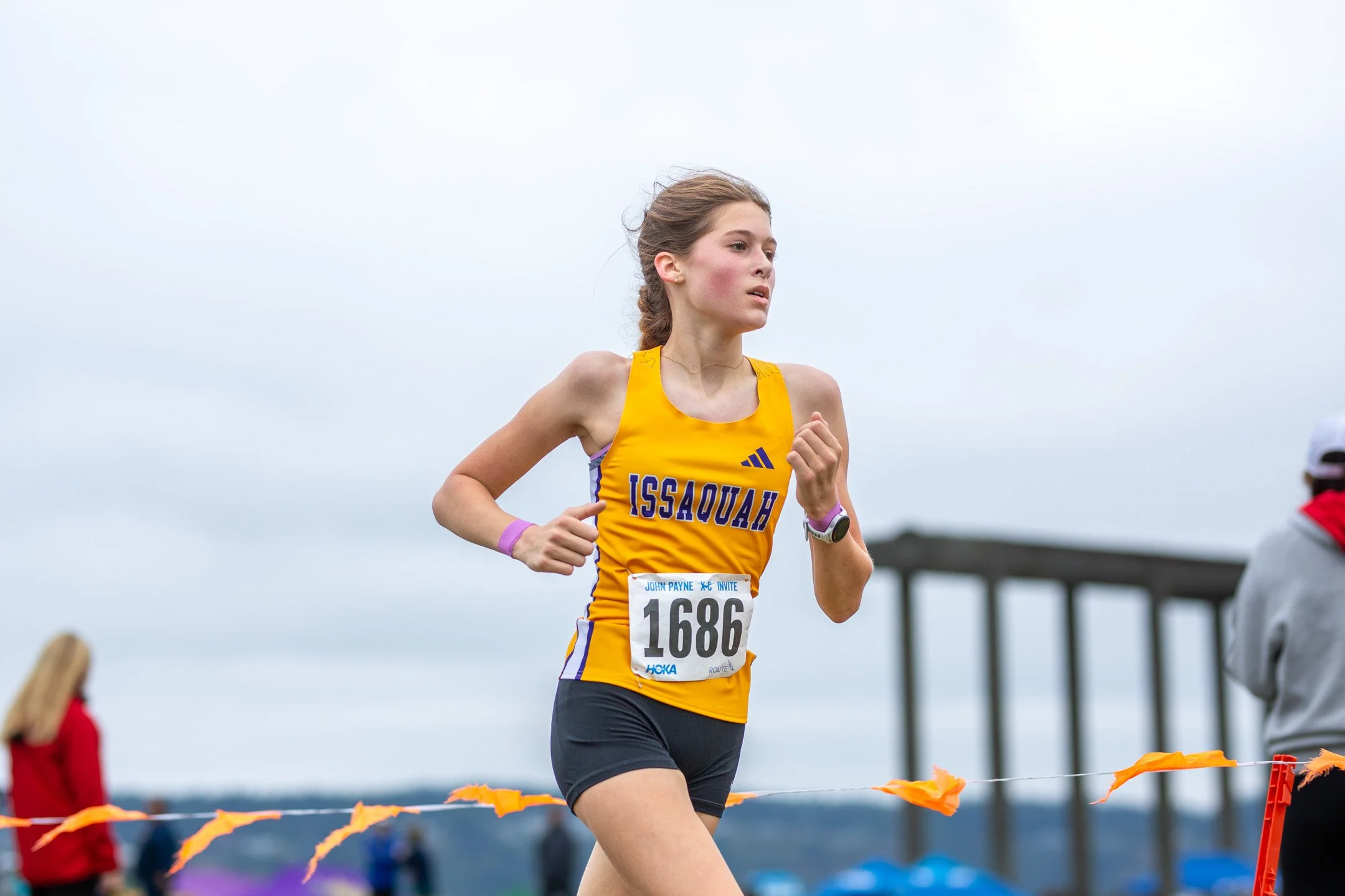 Issaquah High School freshman runner Maya Jaeschke in a gold singlet with bib 1686 running solo along the Chambers Creek course past orange flags.
