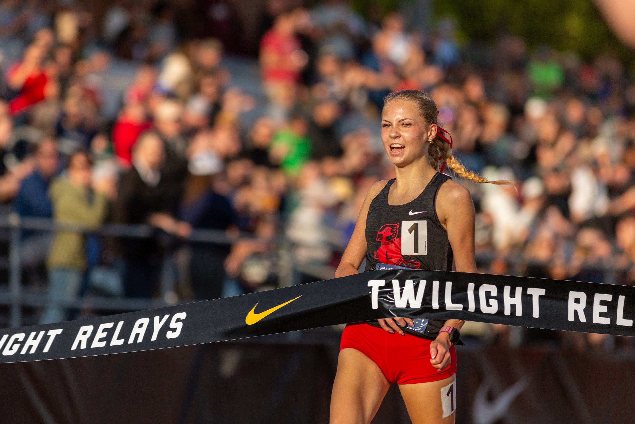 Female track athlete with blonde braid crosses Nike Twilight Relays finish line tape. Smiling runner wears black tank top displaying number one and red shorts during competitive racing event.