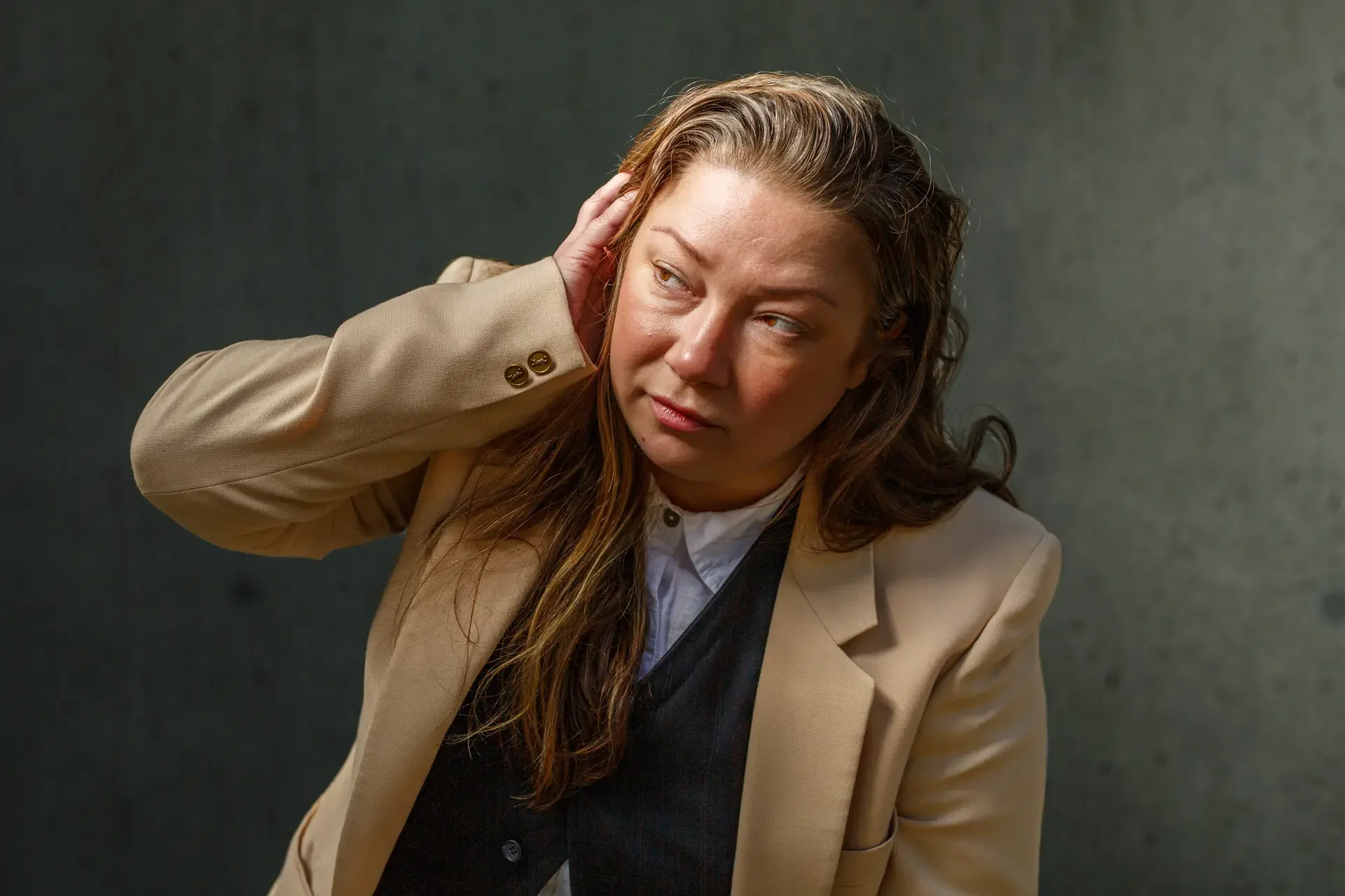 A compressed 85mm portrait of Jennifer Harju wearing a tan blazer and tortoiseshell glasses. She is looking away with a contemplative expression, with her hand raised toward her face against a soft-focus concrete background.