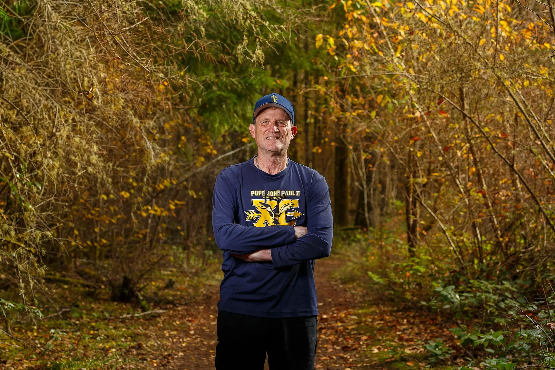 A man in a blue Pope John Paul II shirt and cap stands with arms crossed in an autumn forest.