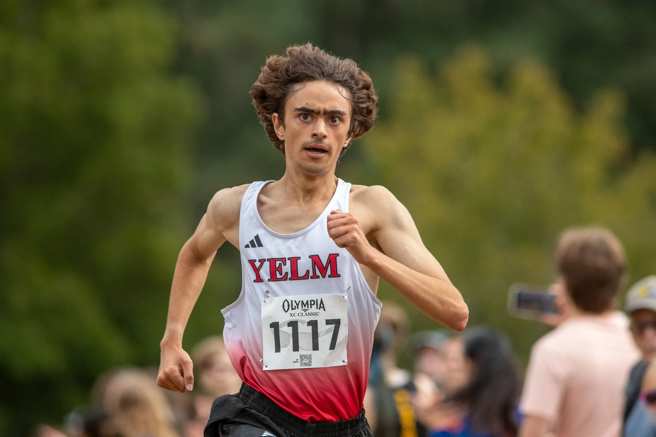 Boy in white and red Yelm jersey, bib 1117, running with a strained expression, spectators visible in the background.