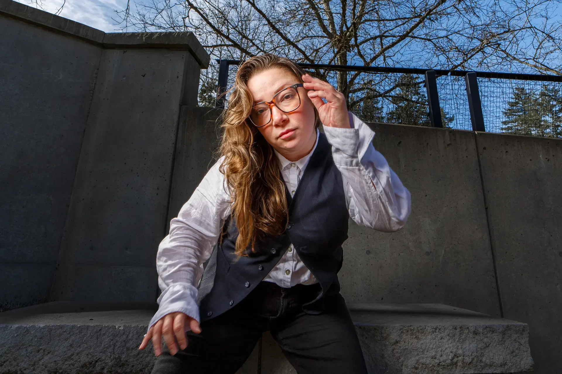 A low-angle shot of Jennifer Harju sitting on concrete bench at the Olympia Capitol Campus, adjusting her glasses and looking directly at the camera. She is wearing a white button-up shirt and pinstripe vest.