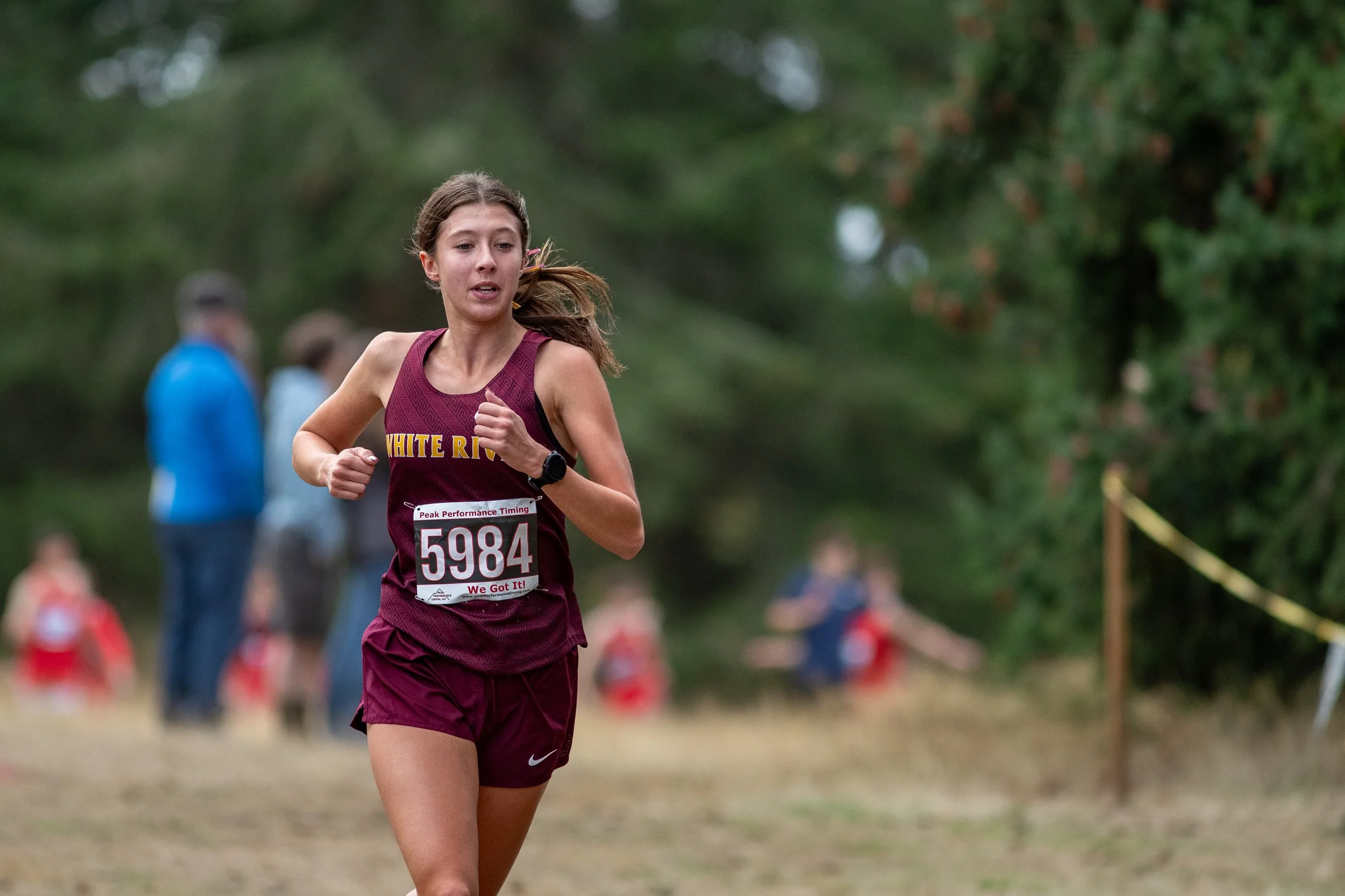 Paeton Poelman of White River, bib 5984, in a maroon singlet and shorts, runs through a dirt clearing with spectators blurred behind.