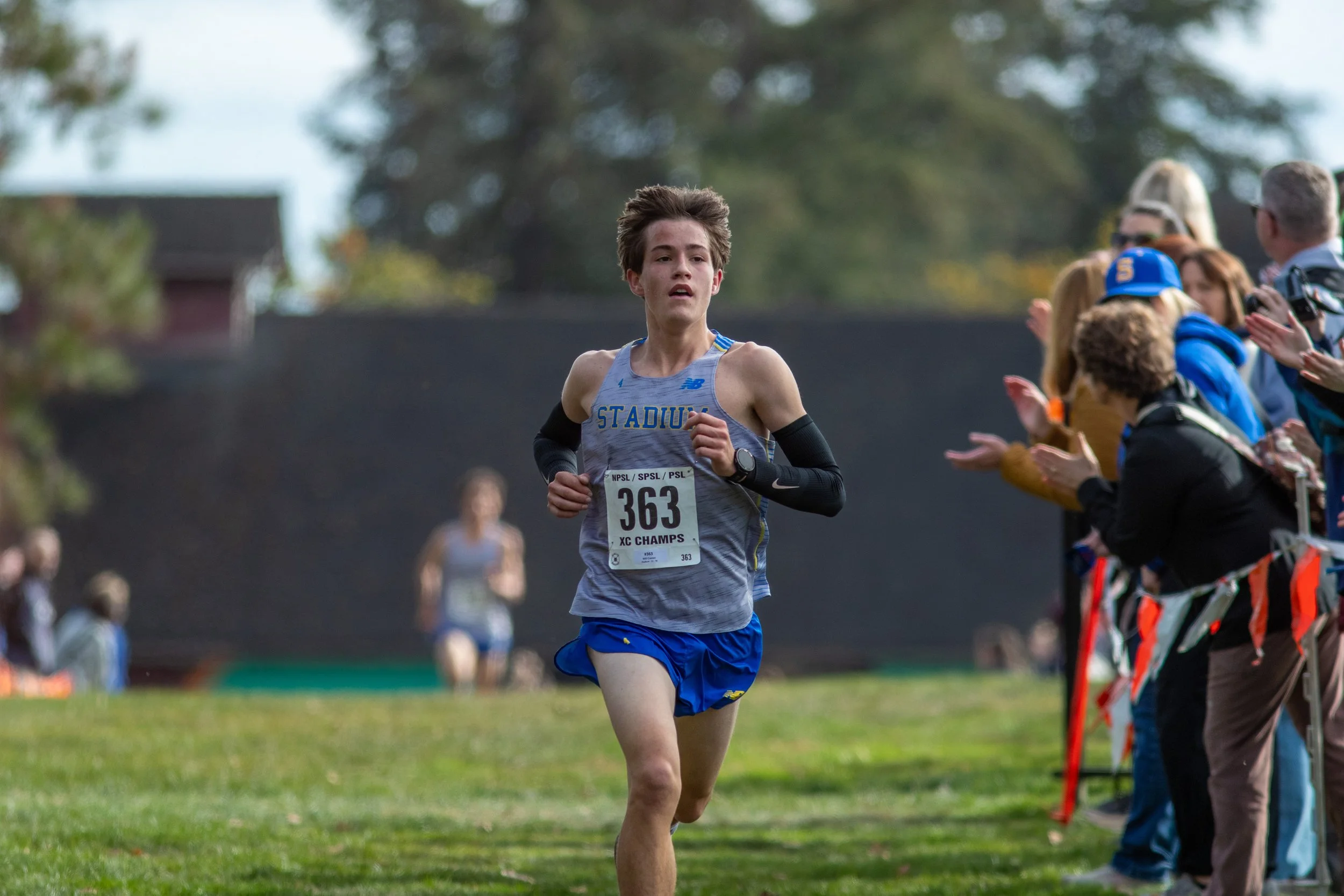 Will Cramer of Stadium, bib 363, runs on grass in a gray Stadium singlet, black arm sleeves and blue shorts, as spectators clap along orange course flags.