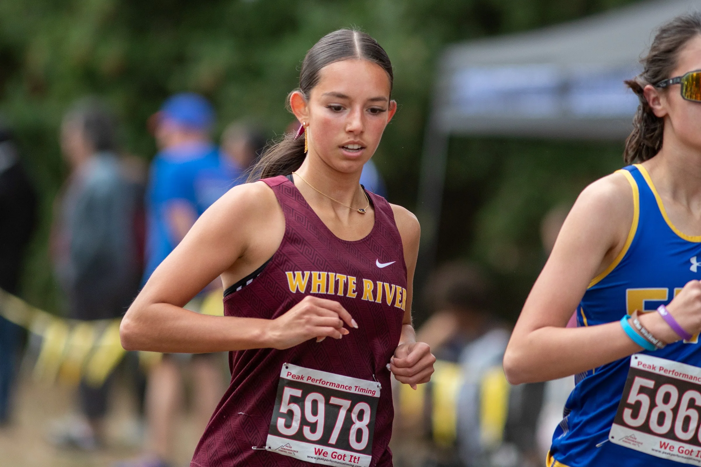White River junior Victoria Blakley, bib 5978, in a maroon White River singlet, running beside a runner in a blue singlet.