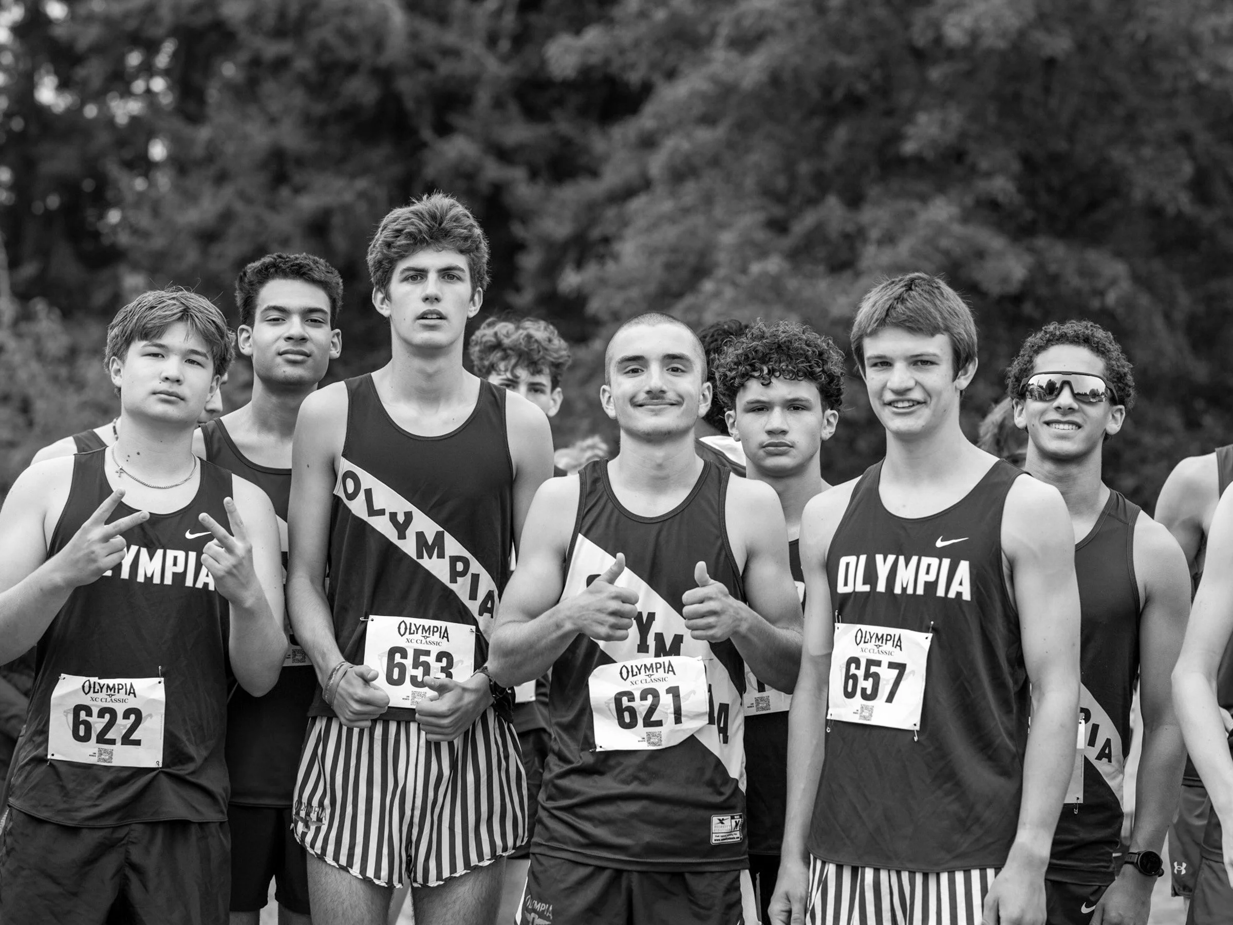 Senior cross country runners in Olympia singlets smiling, giving peace signs and thumbs up before a race.