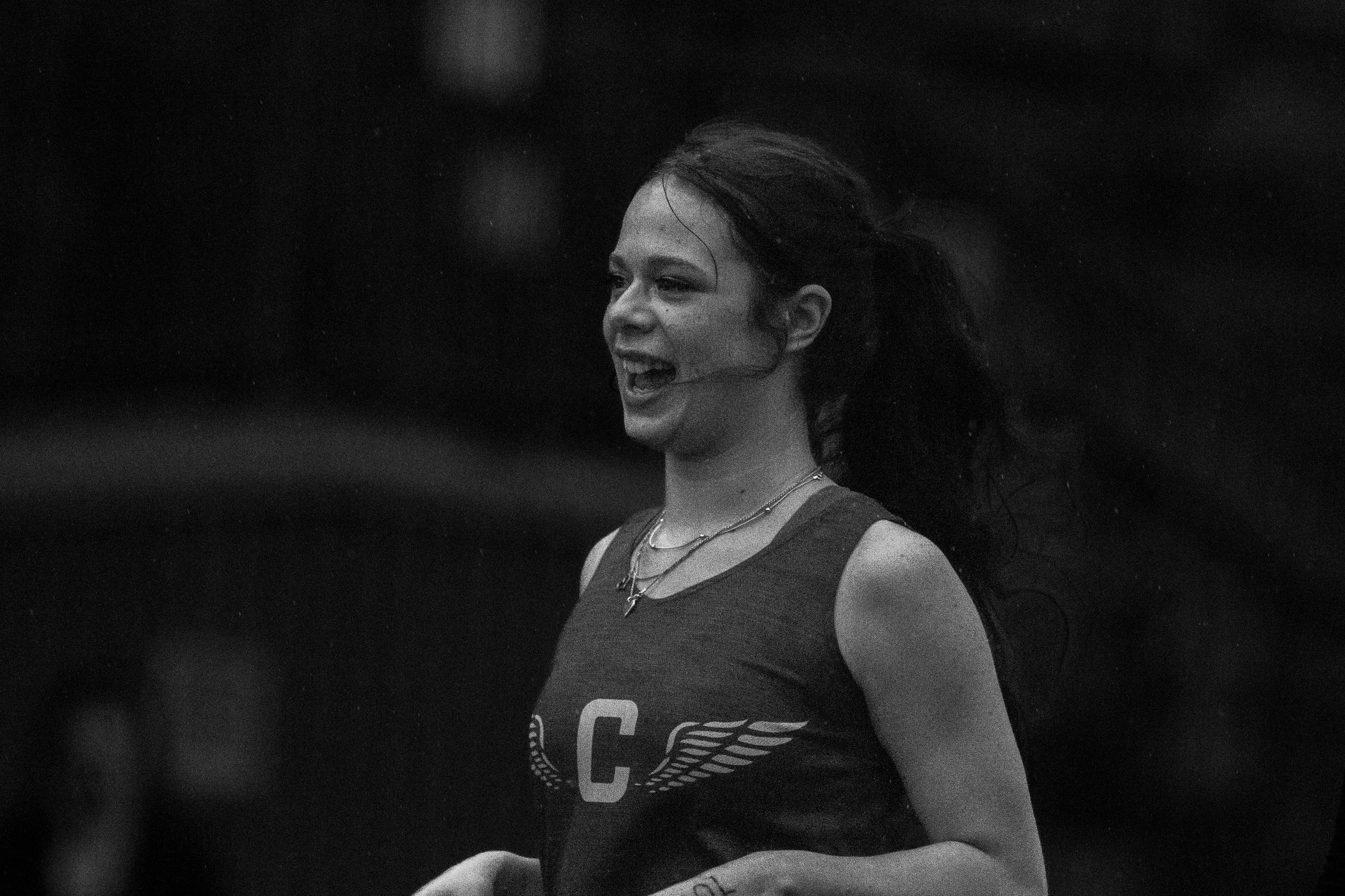 Gritty black and white portrait of a female Capital High School track athlete smiling and laughing in the rain, wearing a dark jersey with a white "C" and wings on the chest.