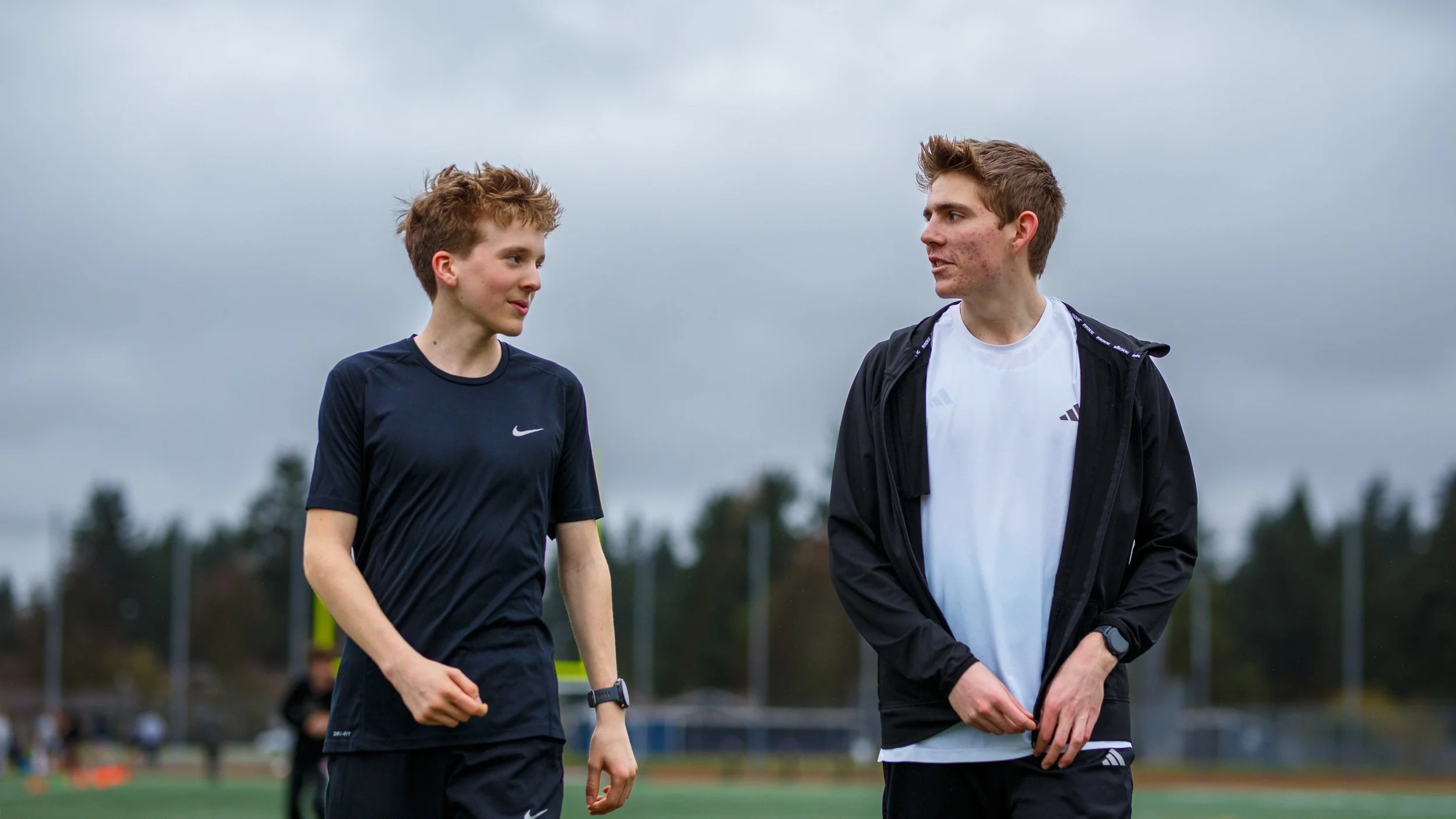 Young male runners talk during training session on outdoor sports field. Athletes wear Nike and Adidas activewear walking on track under overcast sky after completing morning workout.