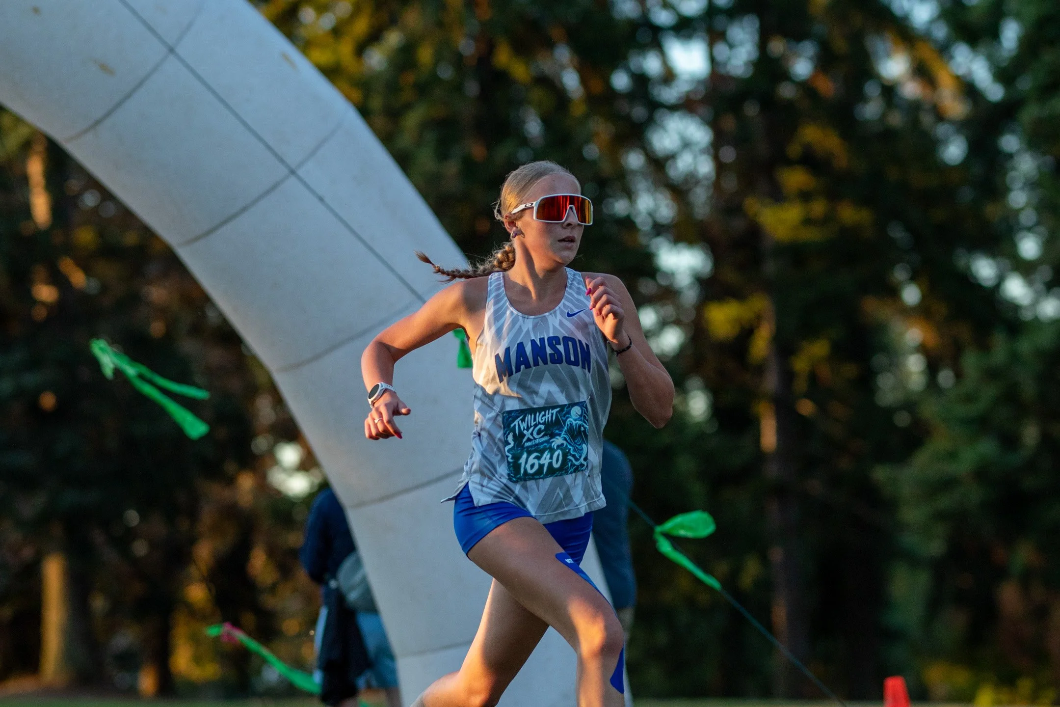 Manson runner Grace Walsh in a white patterned jersey with bib 1640 and sunglasses running past a white inflatable arch in warm golden light.