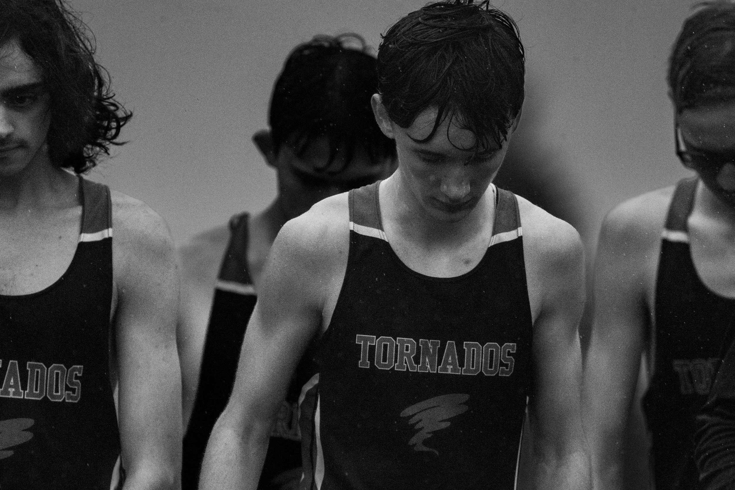 Black and white, heavily grained photograph of three Yelm High School track athletes standing at the starting line. They are wearing dark "Tornados" jerseys with wet hair, looking down with focused expressions as the rain falls.