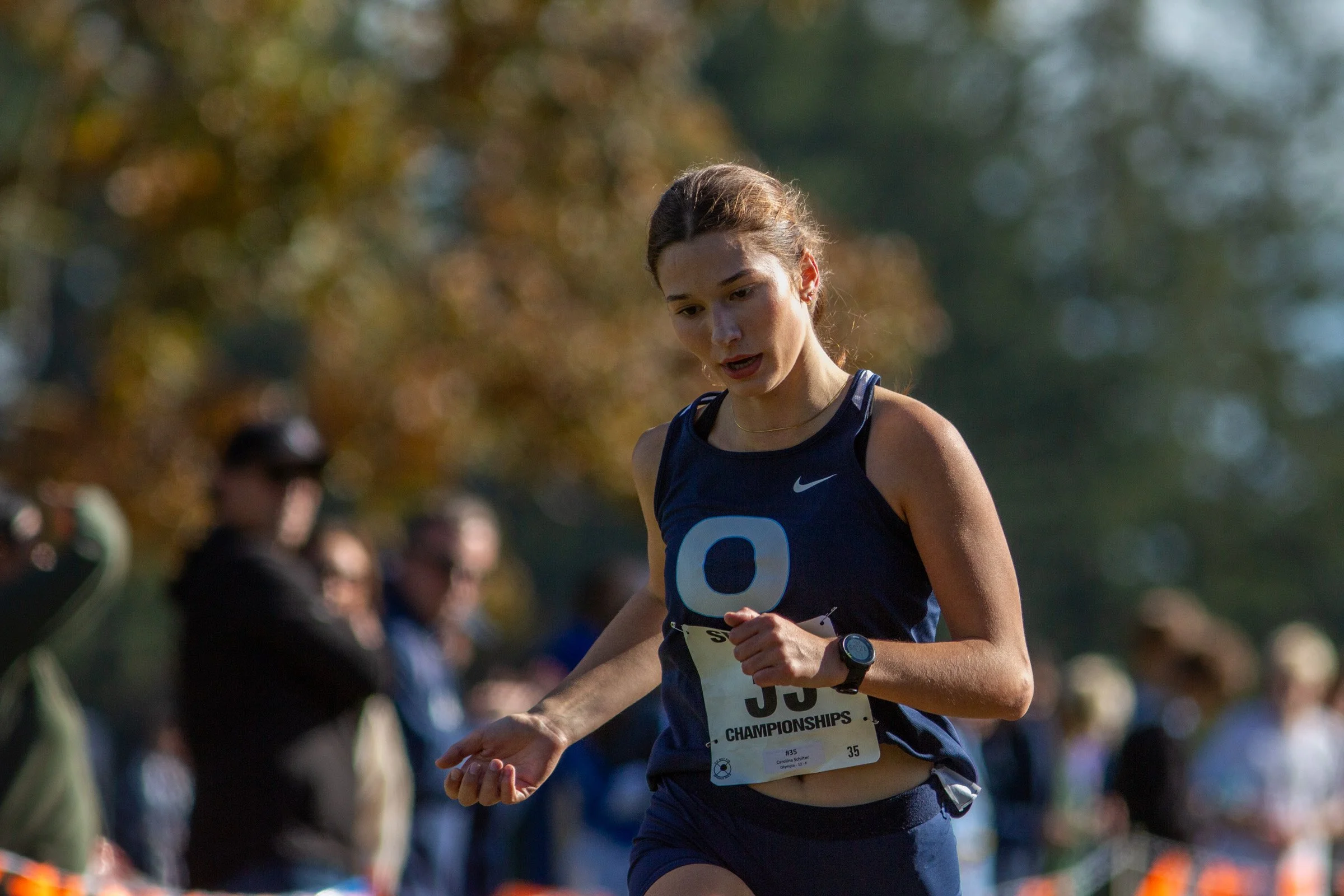 Carolina Schilter of Olympia runs in a navy Olympia singlet with a watch on her left wrist and a championship bib pinned to her front, with blurred spectators behind her.
