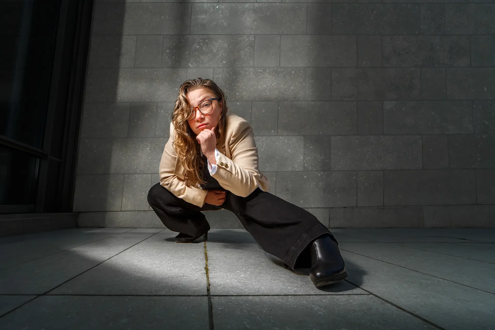 A low-angle environmental portrait of Jennifer Harju in a tan blazer and black wide-leg trousers, performing a dynamic crouched pose against a stark, grid-patterned Brutalist concrete wall.