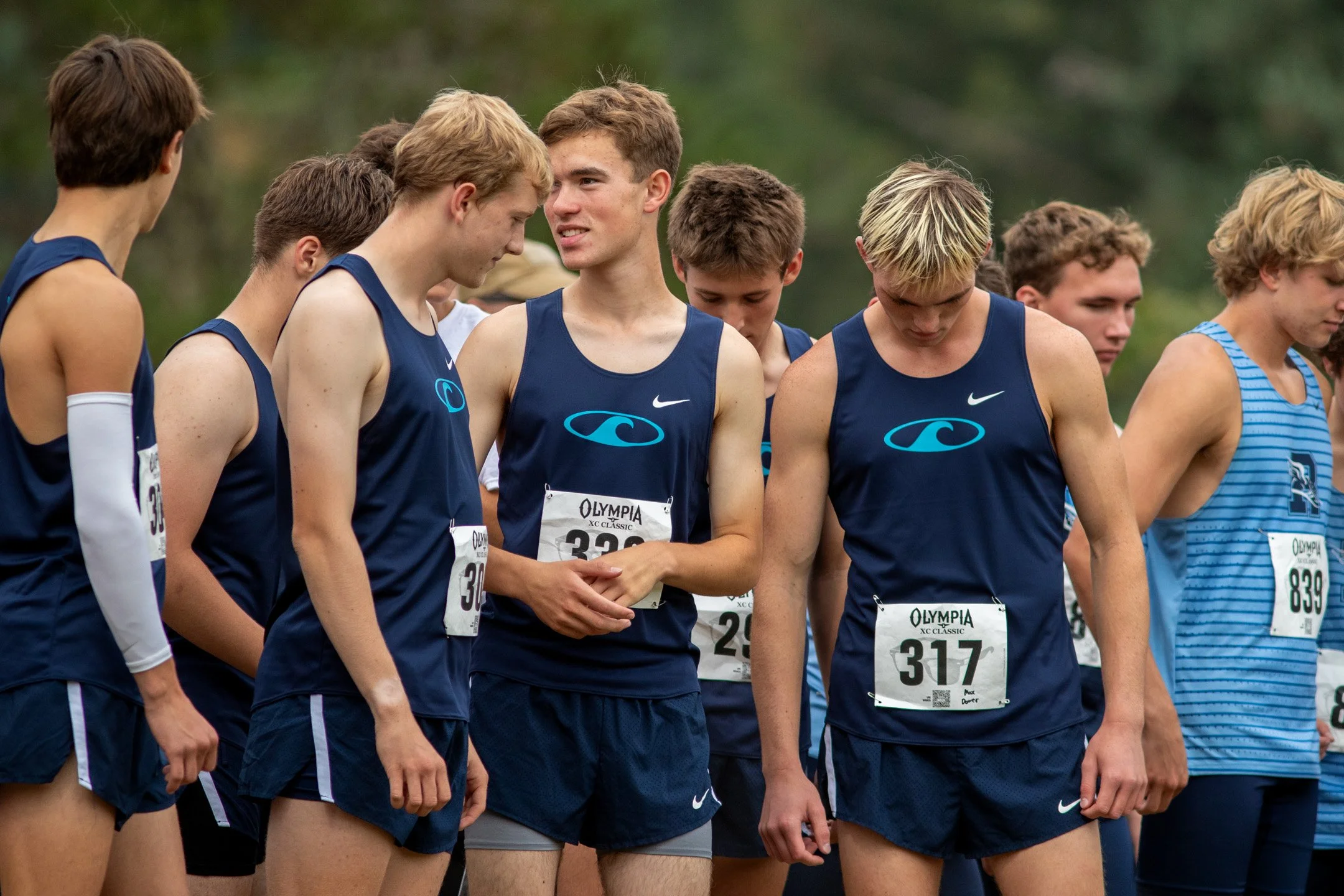 Group of Gig Harbor High School cross country runners in navy jerseys with teal wave logos huddled together before a race, bibs 317 and 839 visible.