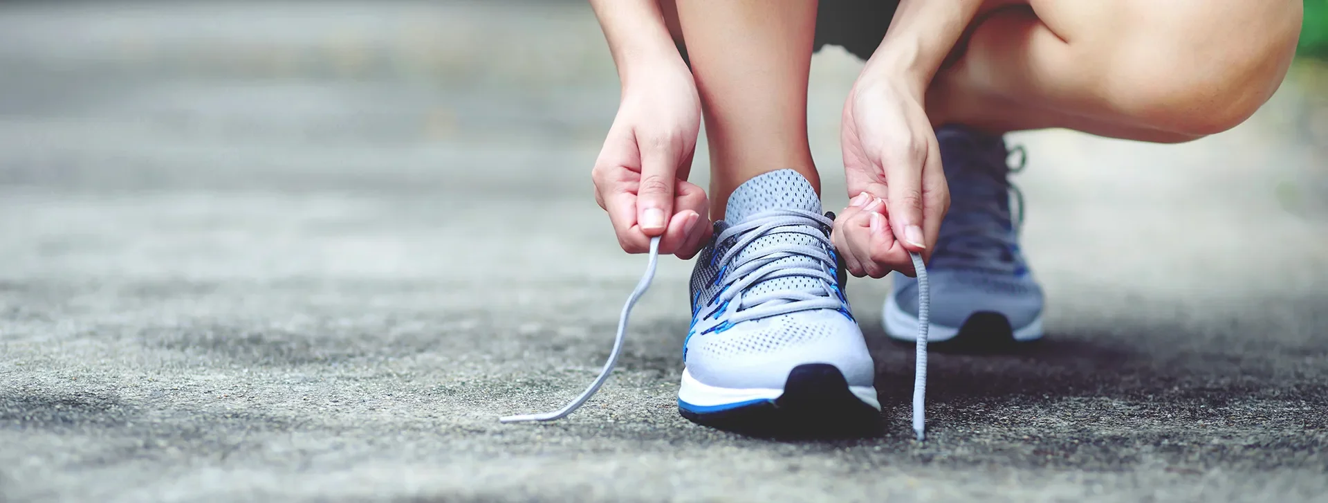 Close-up of unidentifiable hands tying the laces of standard light blue running shoes on a softly blurred asphalt path.