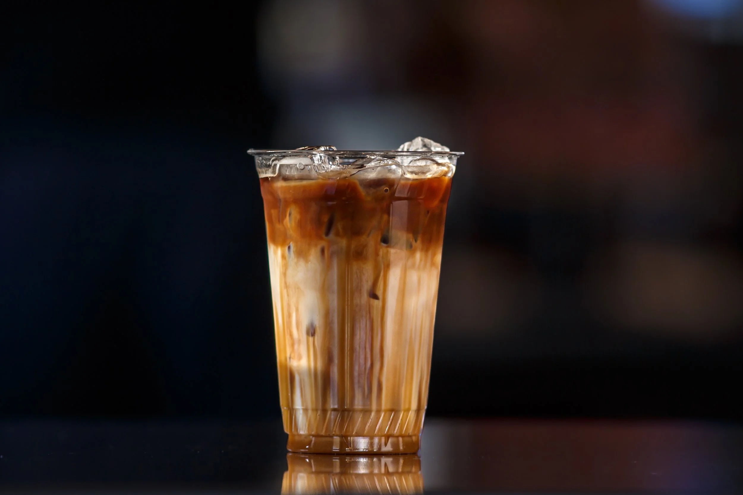 A tall clear plastic cup filled with iced coffee and rich caramel syrup resting on a dark reflective countertop against a moody blurred background at a Hawks Prairie coffee shop.