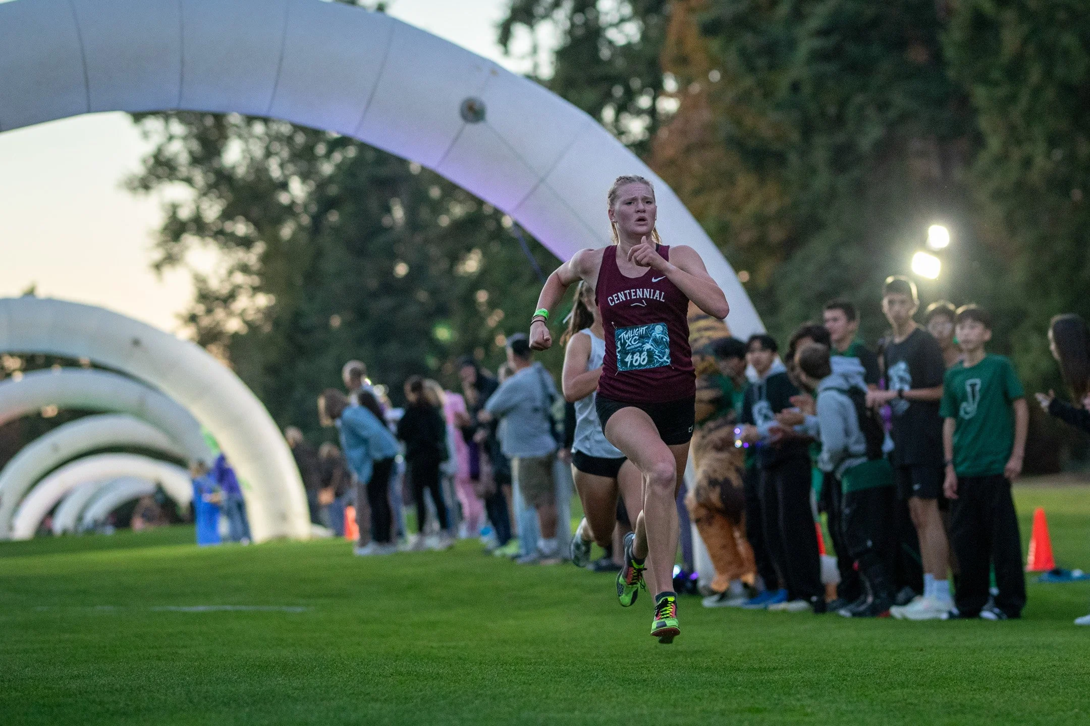 Centennial runner in a maroon jersey with bib 466 sprinting through a tunnel of lighted white inflatable arches in twilight.
