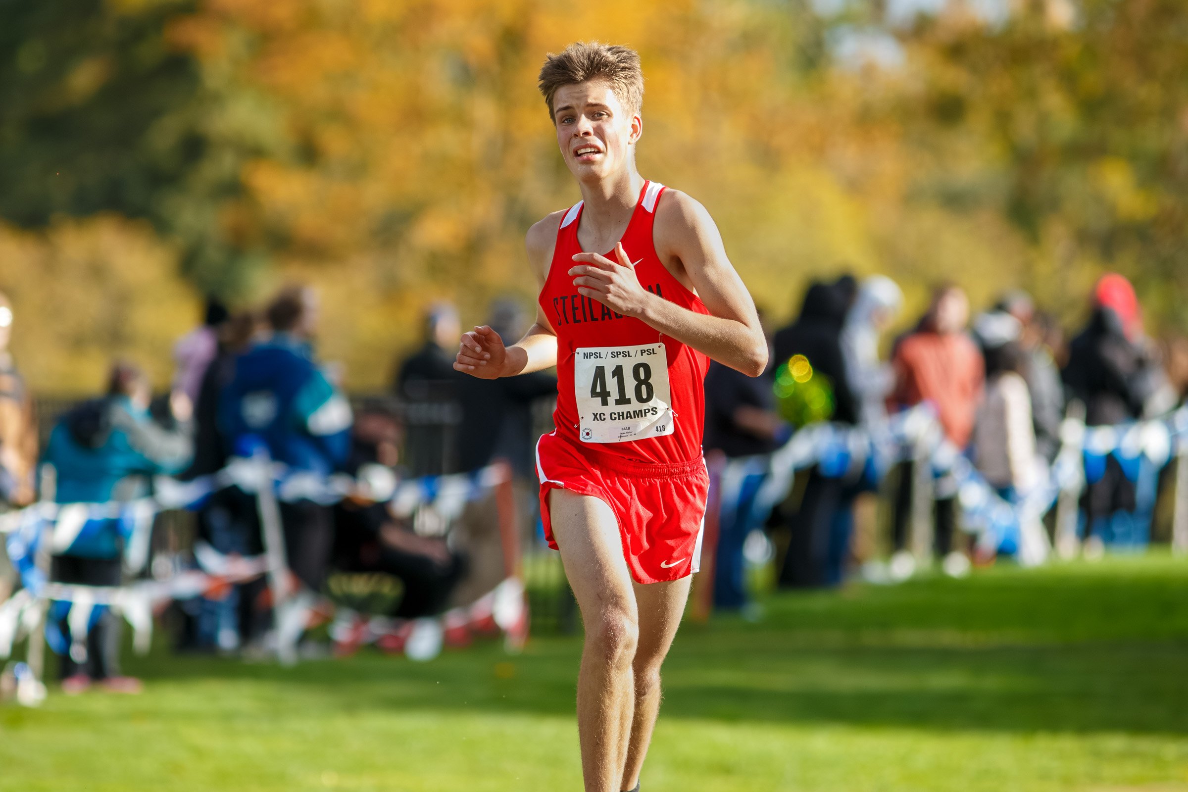 Luke Slater of Steilacoom in a red Steilacoom singlet, bib 418, running toward the camera with fall foliage and spectators lining the course behind him.