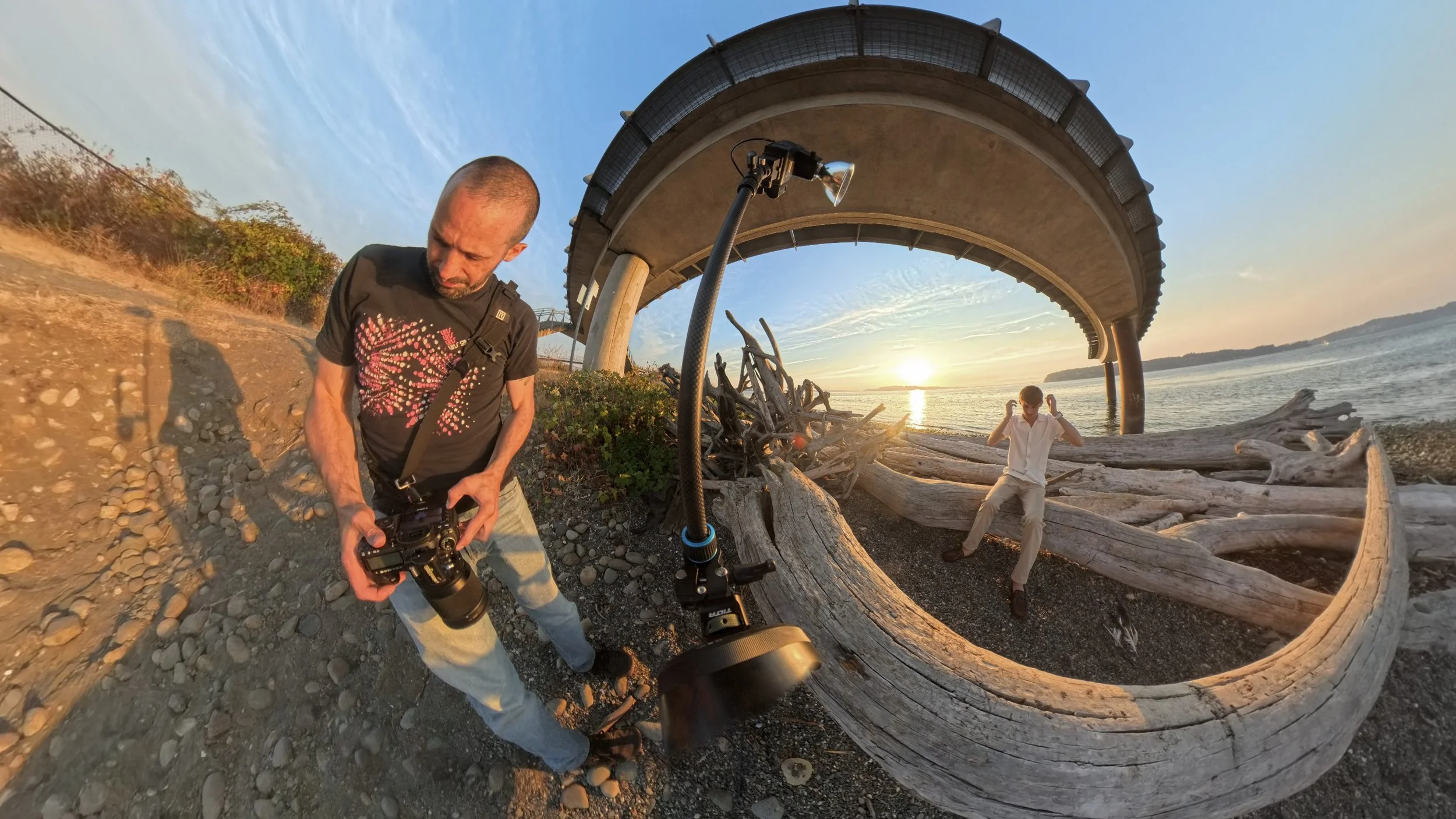 Photographer setting up a Sunpak 120J flash on a light stand at Chambers Bay during a golden hour senior portrait session, subject posed on driftwood in background