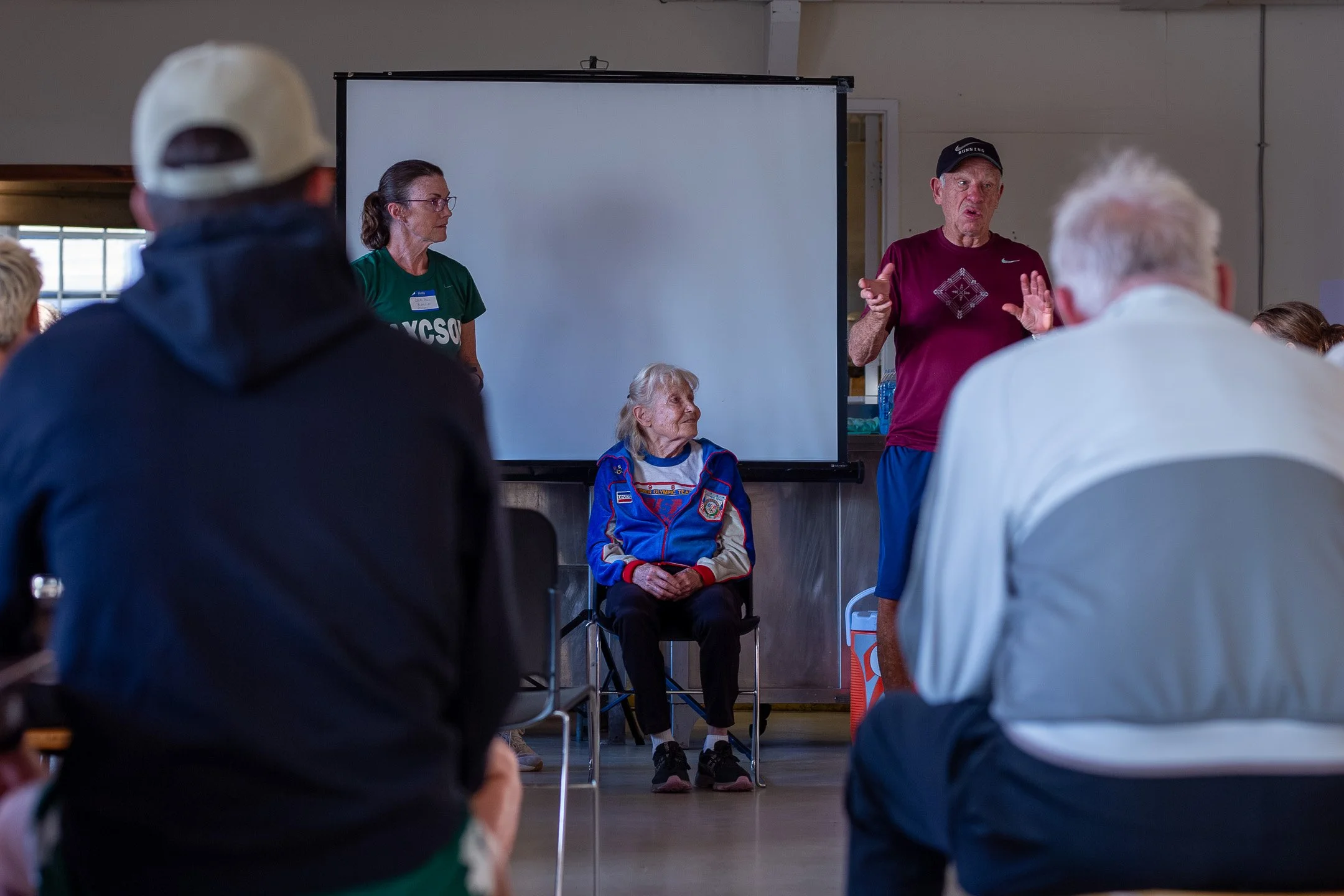 Coaches Pat Tyson and Traci Bianchini standing and conversing with pioneer distance runner Doris Heritage in a meeting hall at Fort Casey during the Whidbey Island Running Camp.