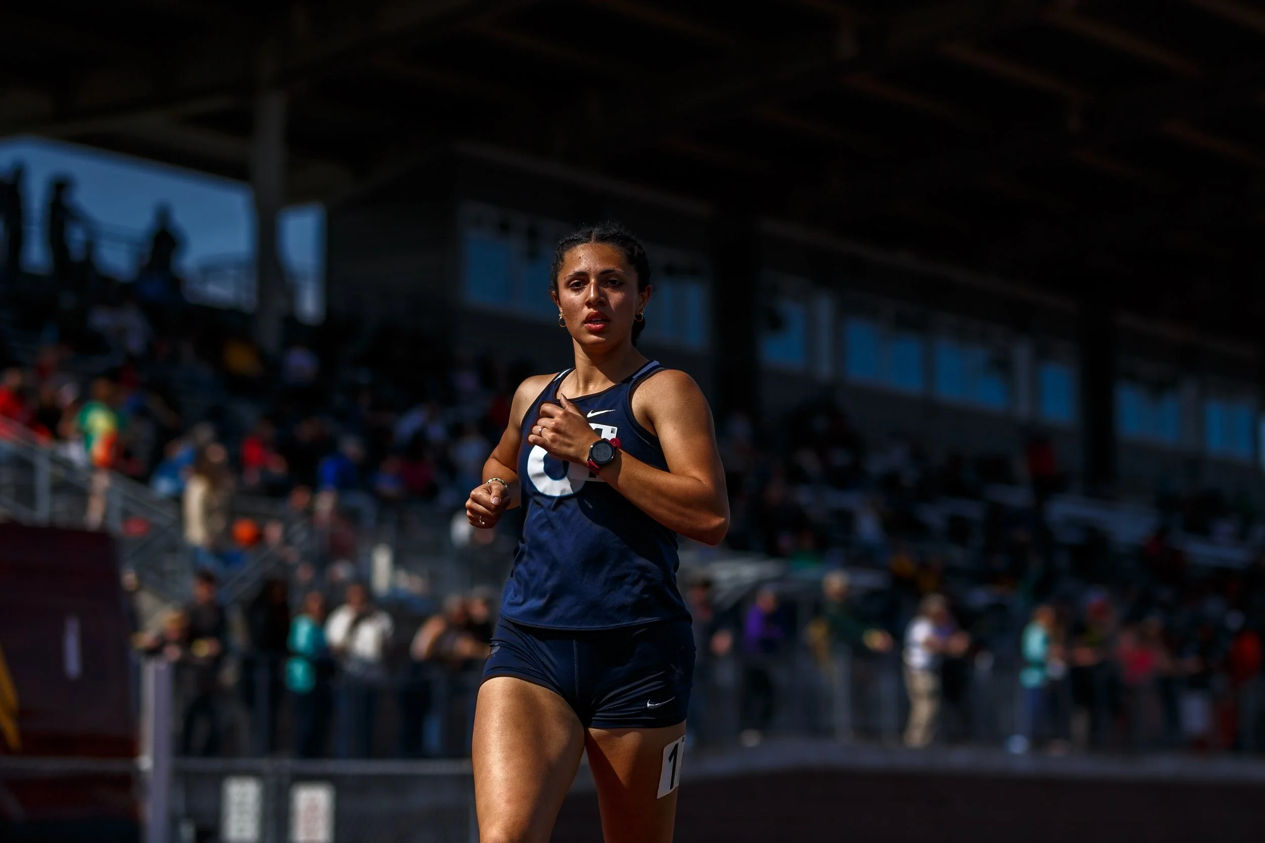 Sofia Dunlap of Olympia High School running at the Mt. Tahoma Track Festival, wearing a navy Olympia singlet, with the crowd in the stands behind her.